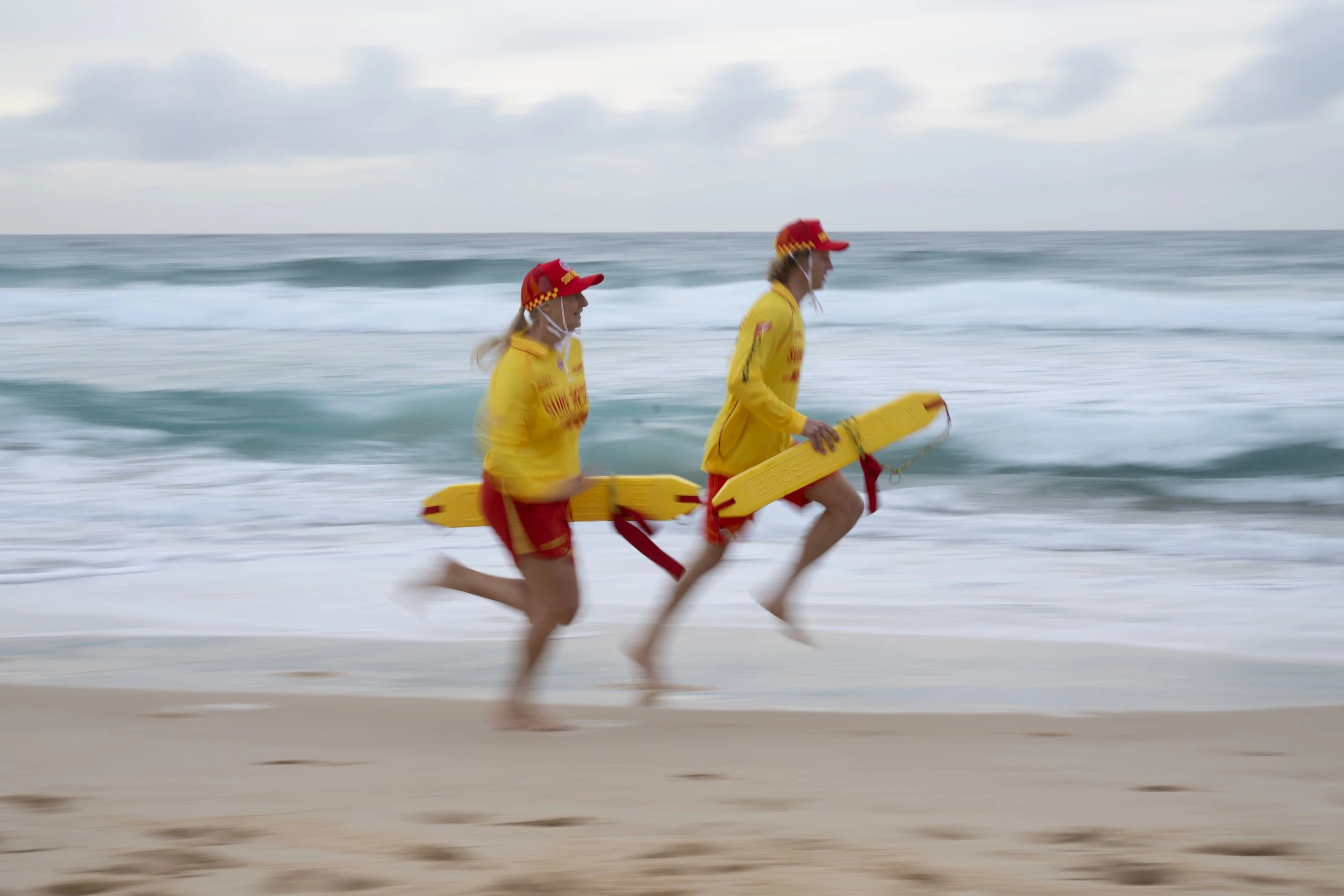 Two lifeguards running on the beach near the water, wearing yellow jackets, red shorts, and red hats, each holding a yellow rescue tube.