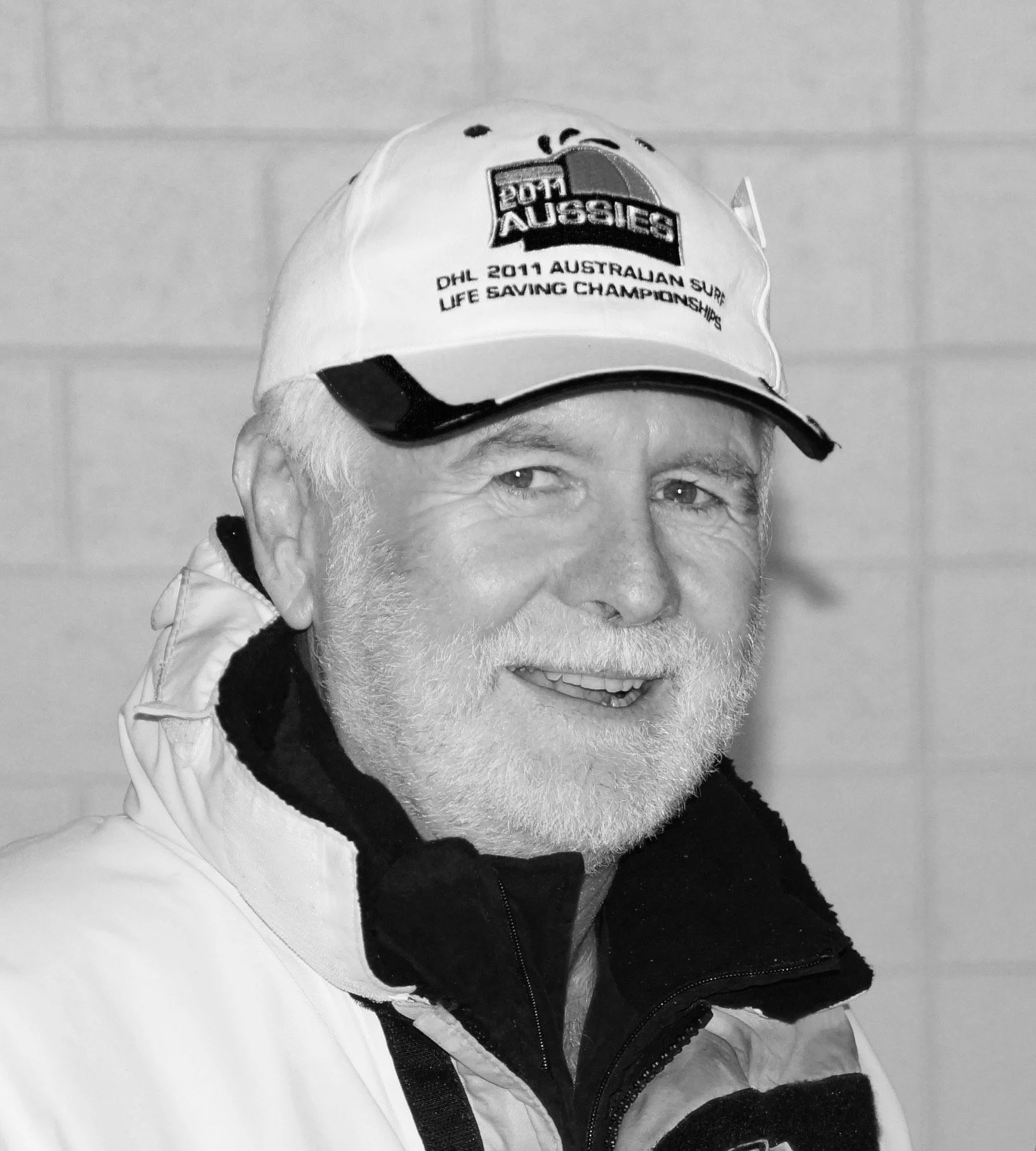 Close-up of an elderly man wearing a white cap with sports logos and text, smiling, with a white and black jacket, in front of a plain background.