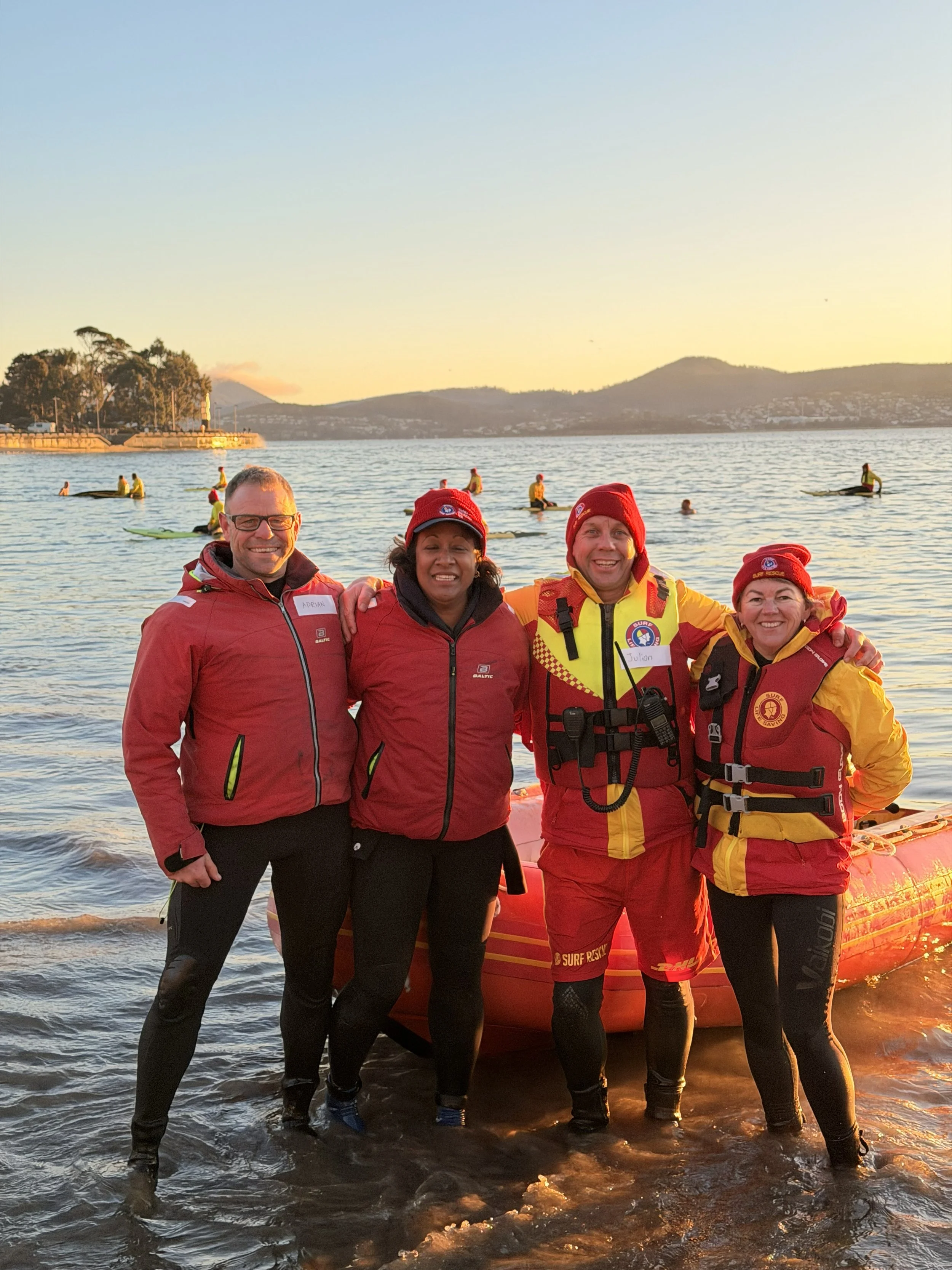 Four rescue workers wearing red and yellow uniforms and life jackets standing in shallow water at a beach during sunset. Behind them, people are on paddleboards in the water, and distant hills and trees are visible.