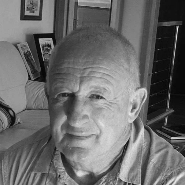 Black and white photo of an older man with short hair, smiling slightly, sitting indoors with framed pictures on the wall behind him.