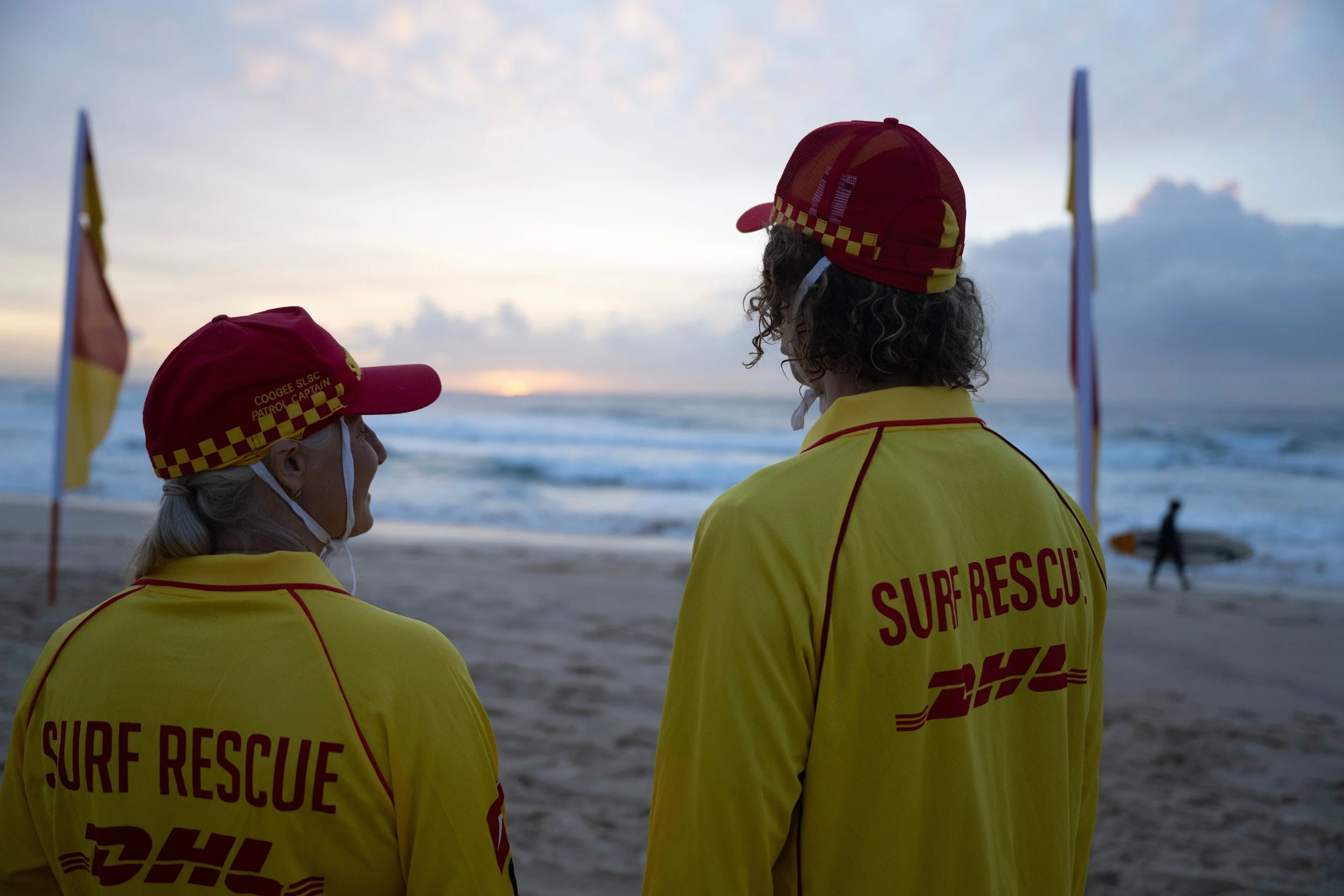 Two surf rescue workers wearing yellow uniforms and red caps with yellow checkered patterns stand on a beach during sunset, facing the ocean, with flags in the background.