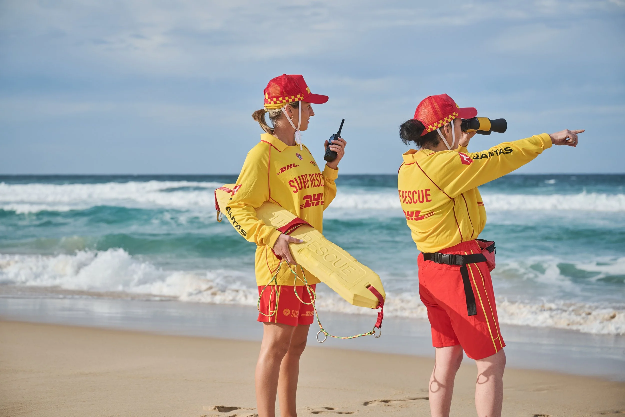 Two female rescue workers in yellow and red uniforms with red hats on a sandy beach. One woman points towards the ocean while the other holds rescue equipment, and the woman with binoculars looks intently at the sea.