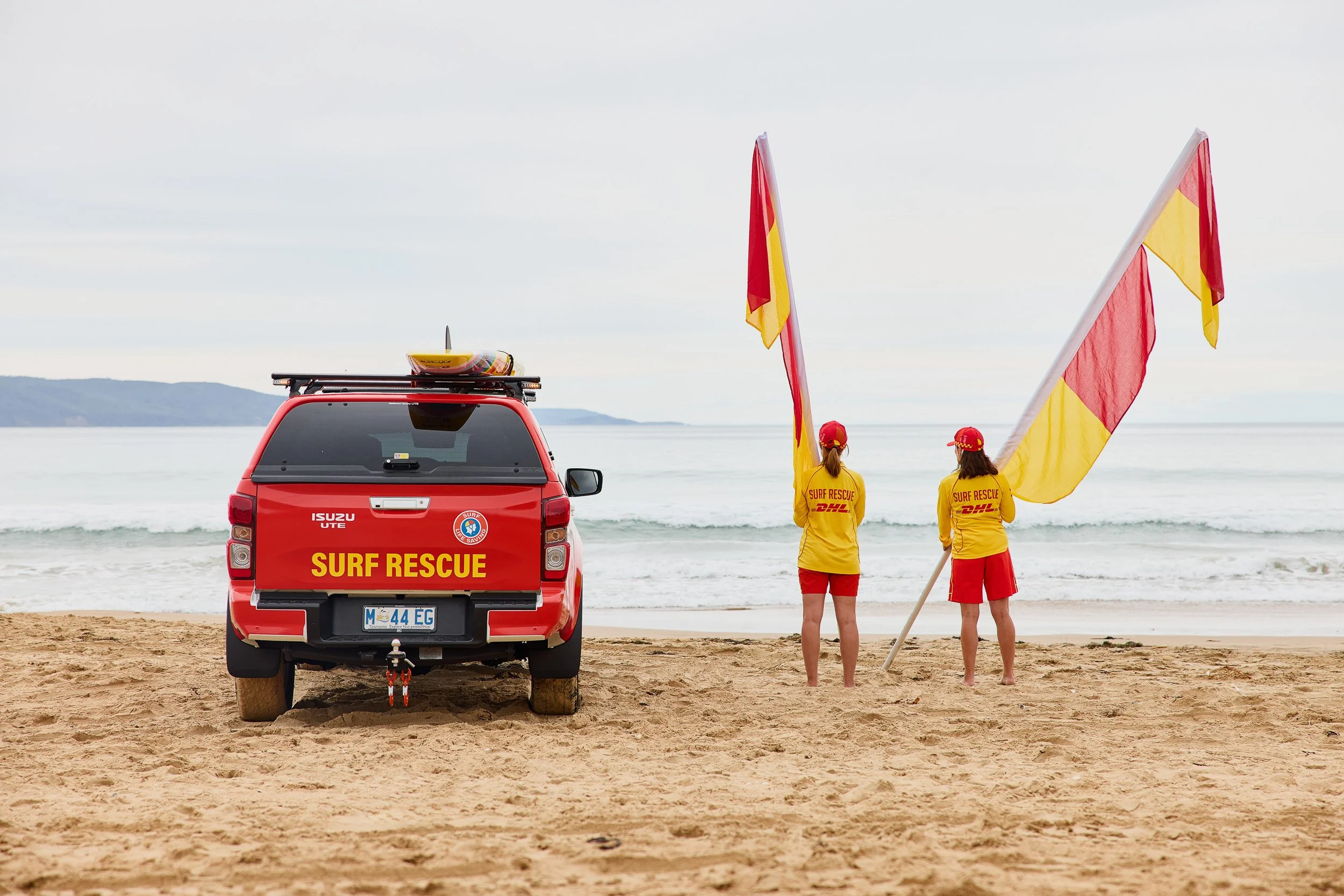 A red surf rescue vehicle parked on a sandy beach with two surf rescue personnel holding yellow and red flags near the shoreline. The personnel are wearing yellow shirts and red shorts, and the scene is overcast with calm sea and distant hills in the background.