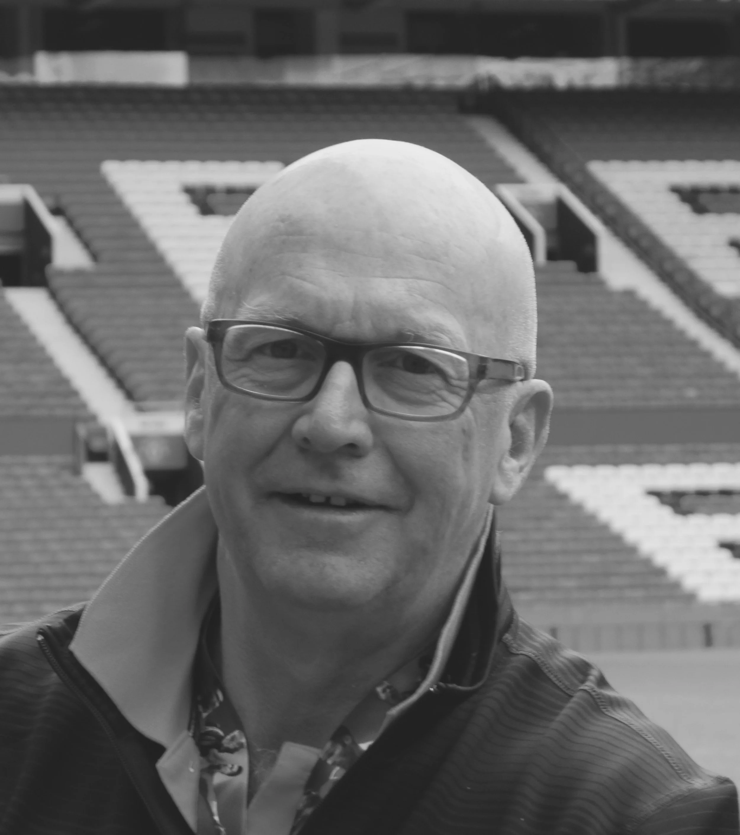 A man with glasses and a bald head in a baseball stadium, with empty stadium seats in the background.