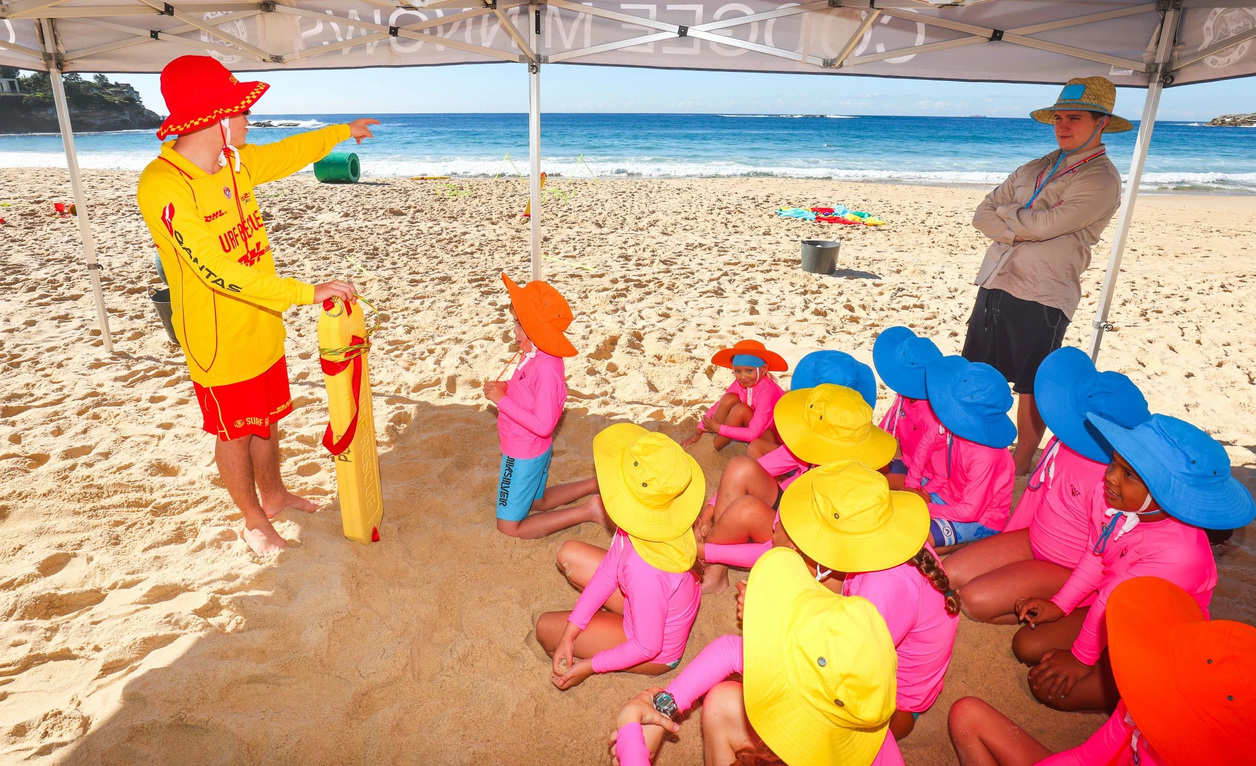 A beach scene showing a group of young children sitting on the sand under a canopy, dressed in pink swimsuits and yellow wide-brimmed hats, listening to a lifeguard. The lifeguard, wearing a yellow and red uniform and a red hat, is standing and pointing towards the ocean while holding a rescue board. An adult man, dressed casually in a beige shirt and hat, stands with arms crossed, watching the scene. In the background, there are waves crashing on the shore and various beach toys scattered on the sand.