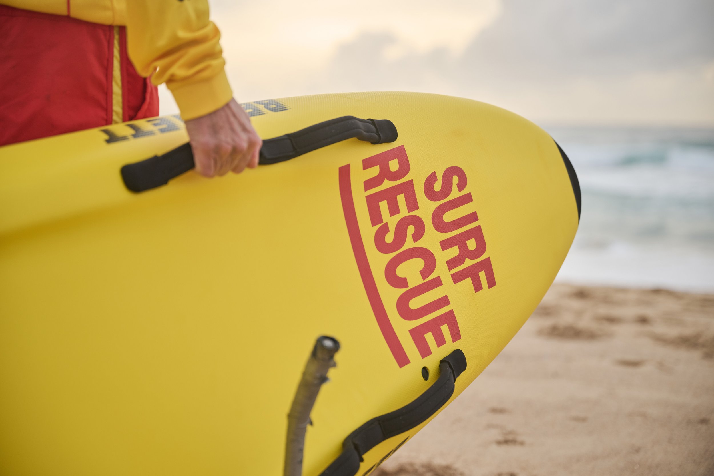 A person holding a yellow rescue surfboard with "Surf Rescue" written on it in red text, standing on a sandy beach with ocean waves in the background.