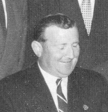 Black and white photo of a smiling man in a suit and tie, with glasses