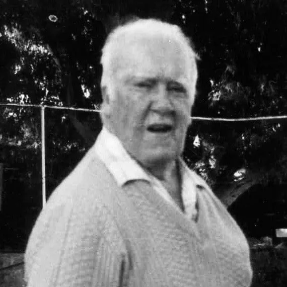 Black and white photo of an elderly man with short white hair, wearing a collared shirt and a sweater, standing outdoors with trees in the background.