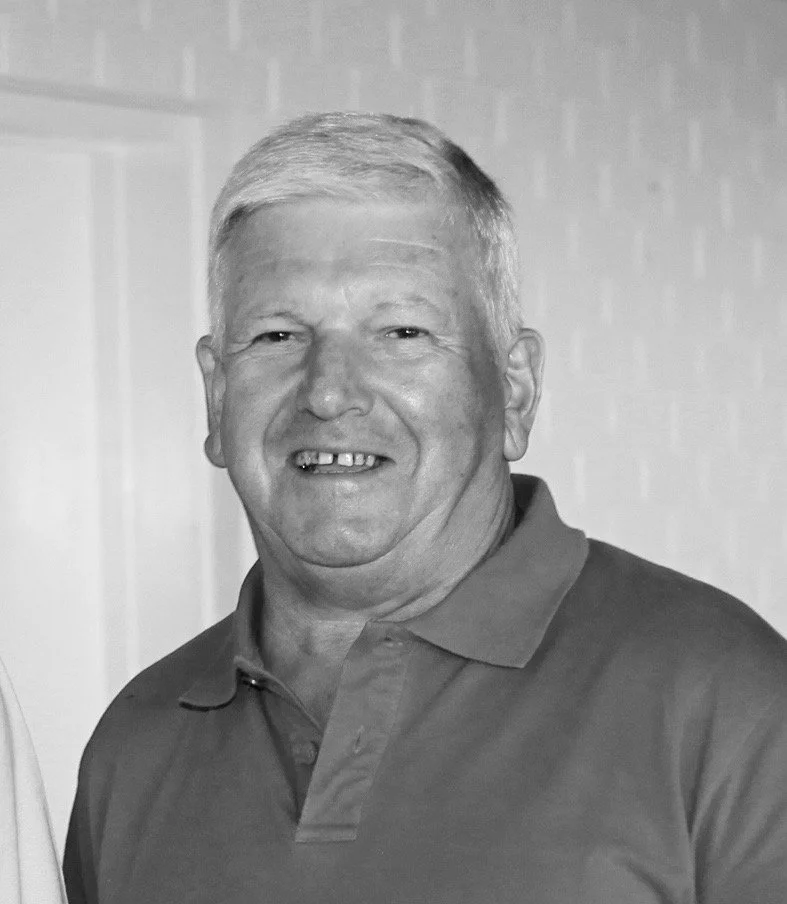 A smiling middle-aged man with short gray hair, wearing a polo shirt, standing indoors against a light-colored wall.
