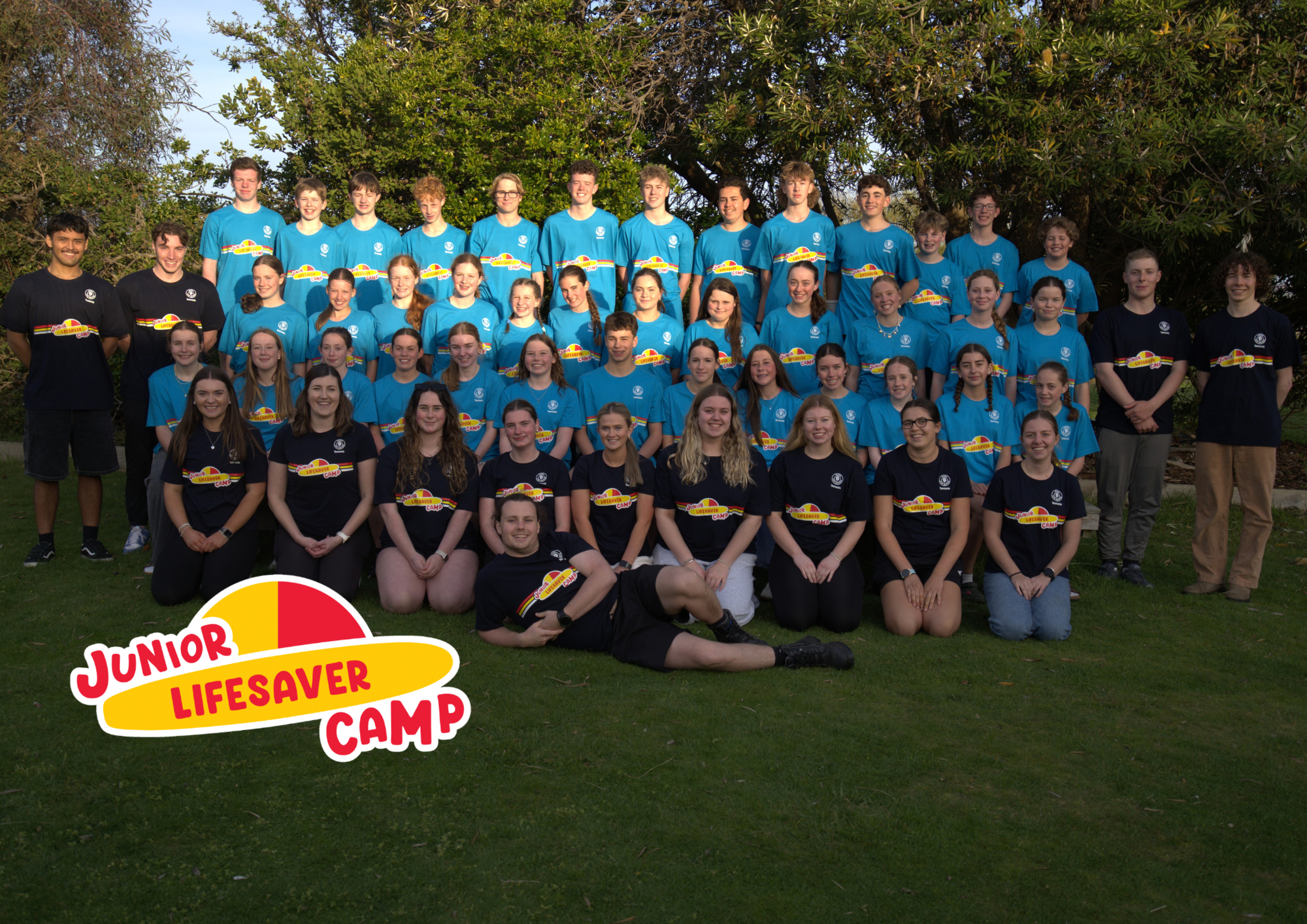 Group of children and teenagers, posing outdoors in front of trees, wearing matching blue and black T-shirts with 'Junior Lifesaver Camp' logo, smiling for a group photo.