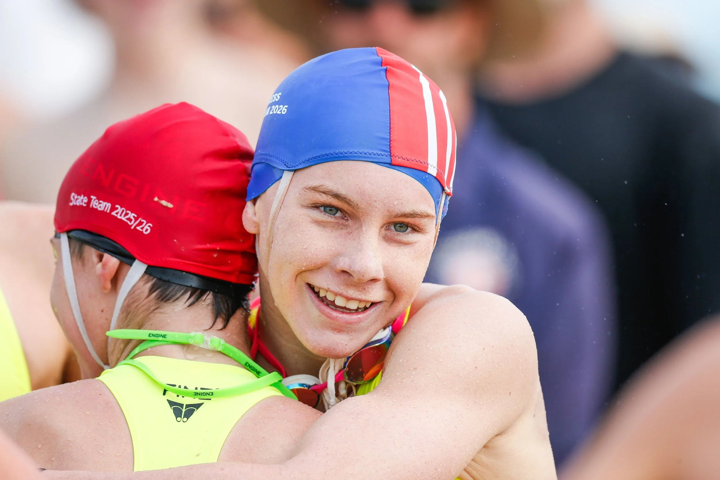 Two competitive swimmers hugging, smiling with water droplets on their skin. The swimmer in front has blond hair, blue eyes, and is wearing a blue, red, and white swim cap. The other swimmer, partly visible, wears a red swim cap with white text and bright yellow-green goggles.