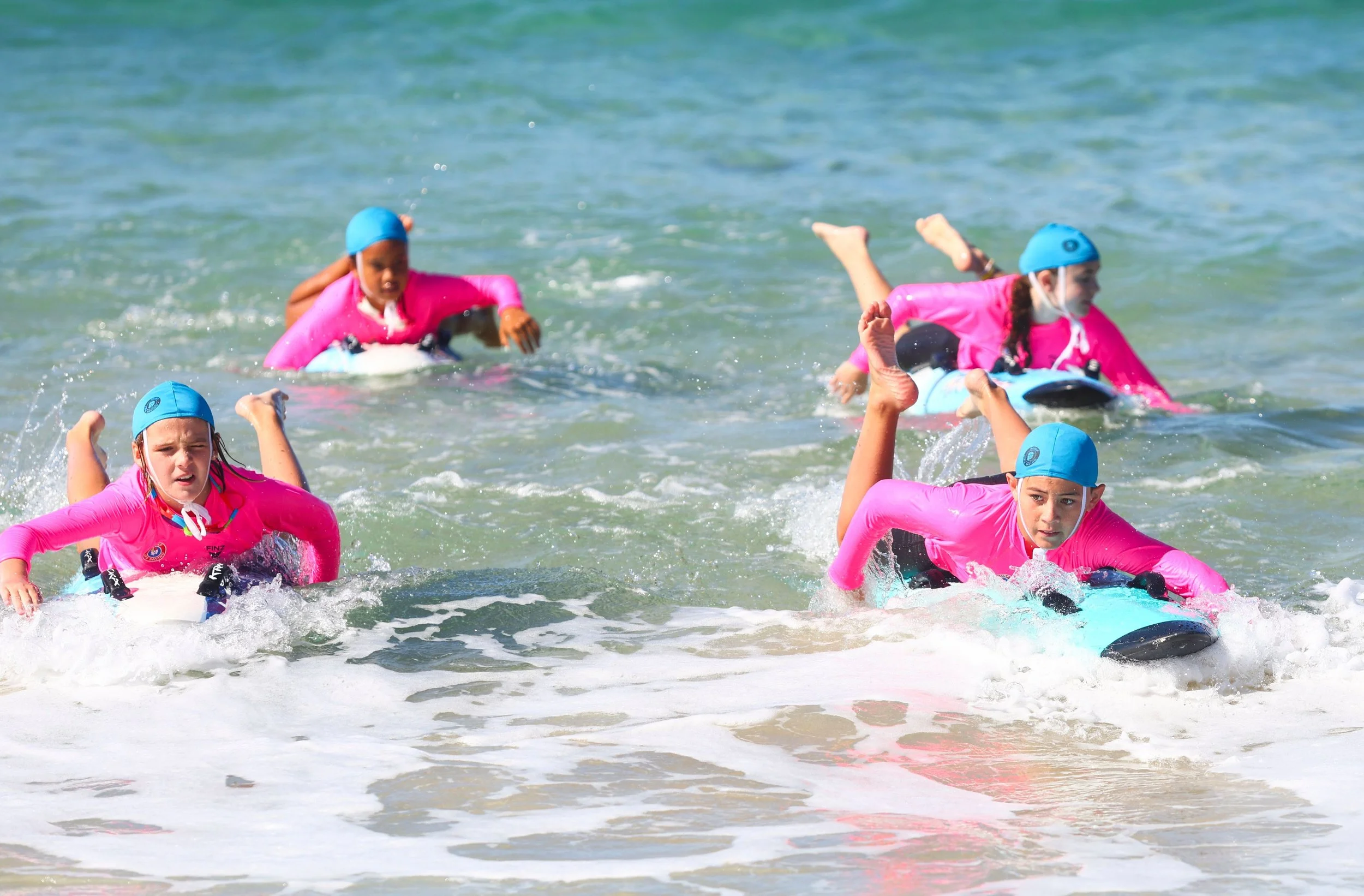 Children in pink swimwear and blue swim caps riding surfboards in the ocean, with some falling off and the waves splashing.