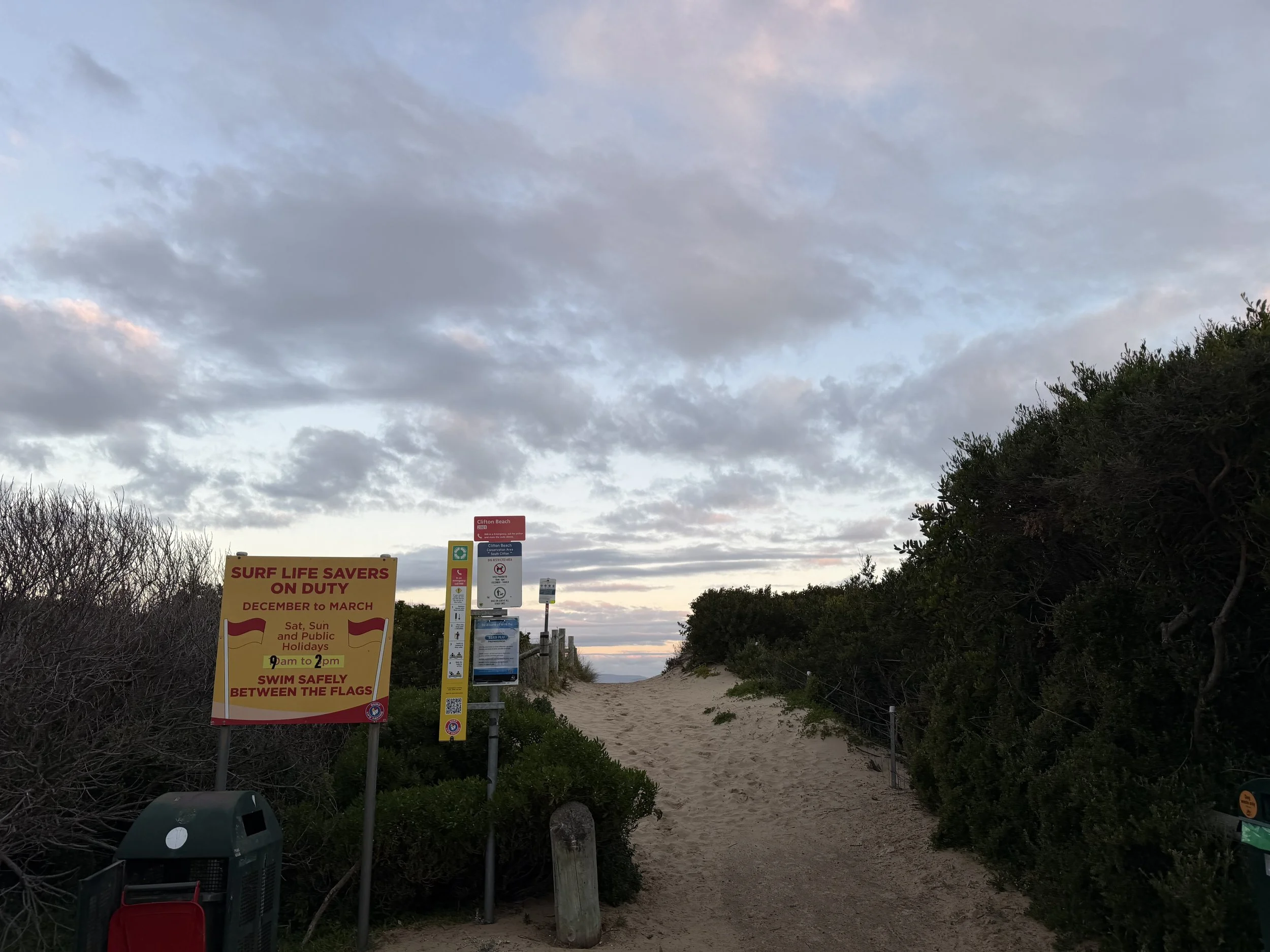 A tall yellow safety and emergency information sign for Park Beach, with instructions on how to use the rescue tube and what to do in an emergency, located outdoors near a body of water.