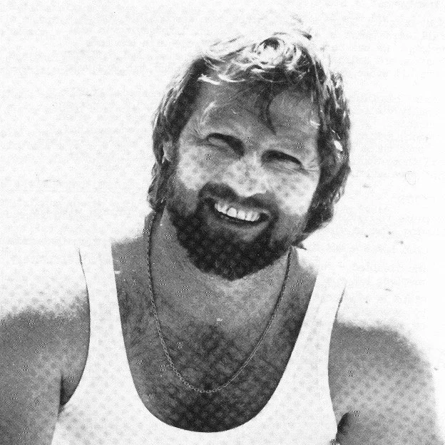 A smiling man with a beard and curly hair, wearing a sleeveless shirt and multiple necklaces, in black and white.