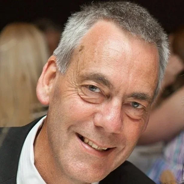A middle-aged man with short gray hair, blue eyes, and a cheerful smile, wearing a dark suit and white shirt, in an indoor setting.