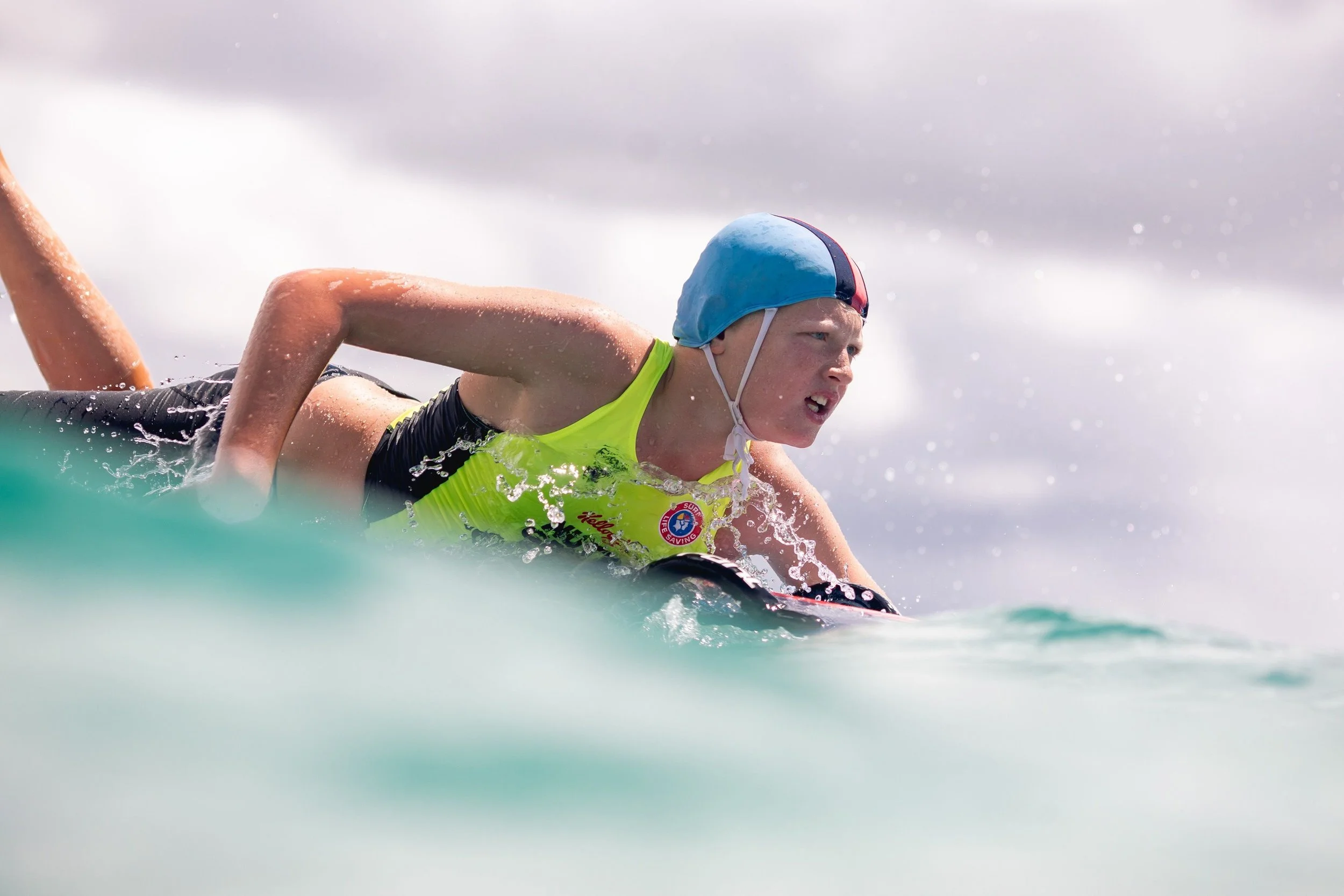 A young swimmer in a yellow swim shirt and blue swim cap practicing open water swimming in rough waters, with a cloudy sky in the background.