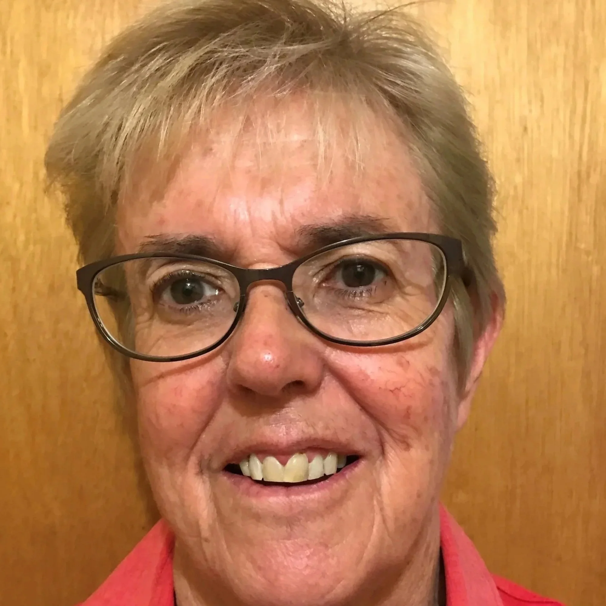 Close-up of an elderly woman's face, showing her glasses, brown eyes, and smiling mouth, with short blonde hair and a red shirt.