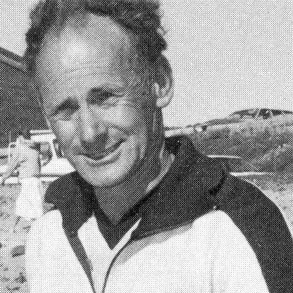 Black and white photo of a smiling man with curly hair, wearing a jacket, outdoors with land and harbor in the background.