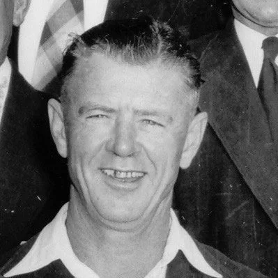 Black and white photo of a smiling man with short hair, wearing a collared shirt and jacket.