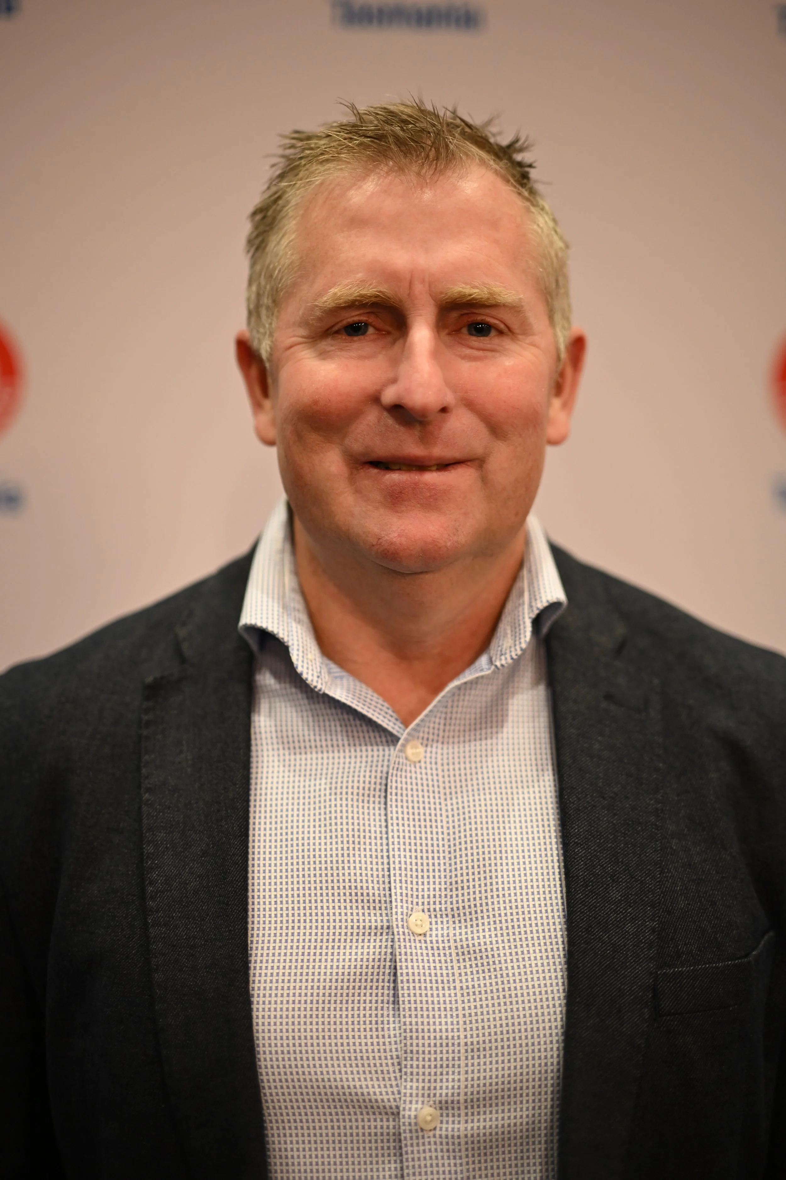 A middle-aged man with short, light-colored hair, wearing a dark blazer and a light-colored, patterned dress shirt, standing against a neutral background.