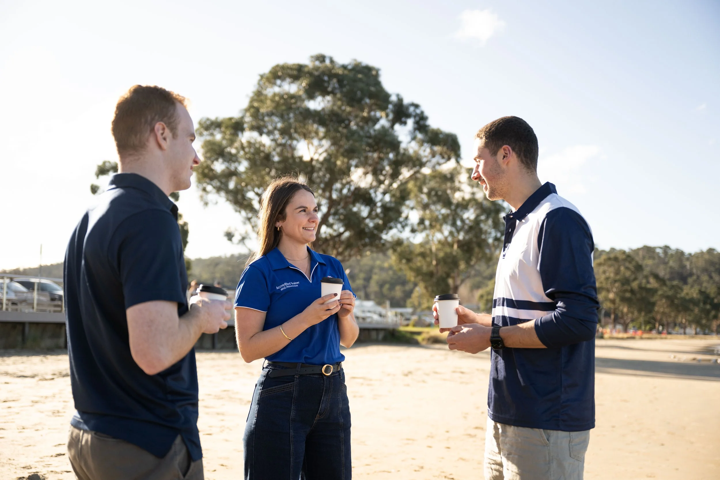 Three people standing outdoors, engaged in conversation, with trees and a parking lot visible in the background.