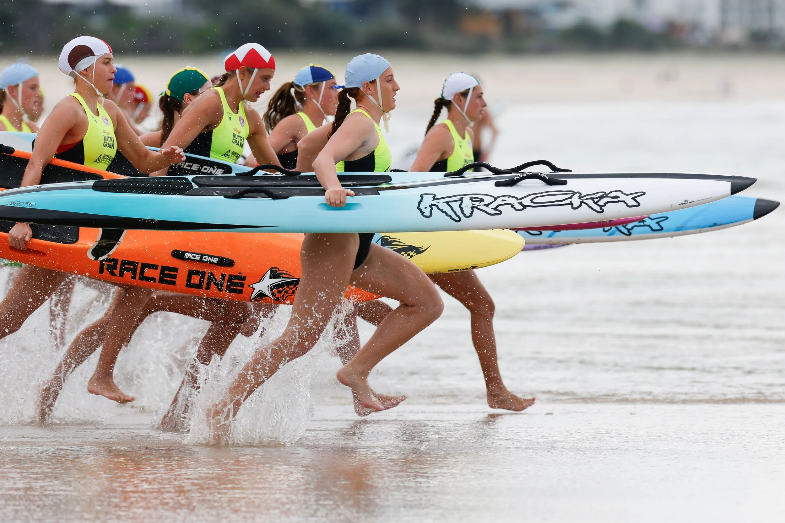 Group of female athletes wearing life saver caps and yellow racing uniforms, running barefoot in water while holding paddles, participating in a kayak race on a beach.