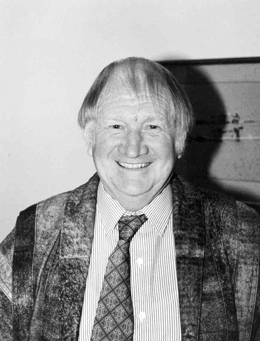 Black and white photo of a man smiling, wearing a patterned tie, a collared shirt, and a textured jacket.