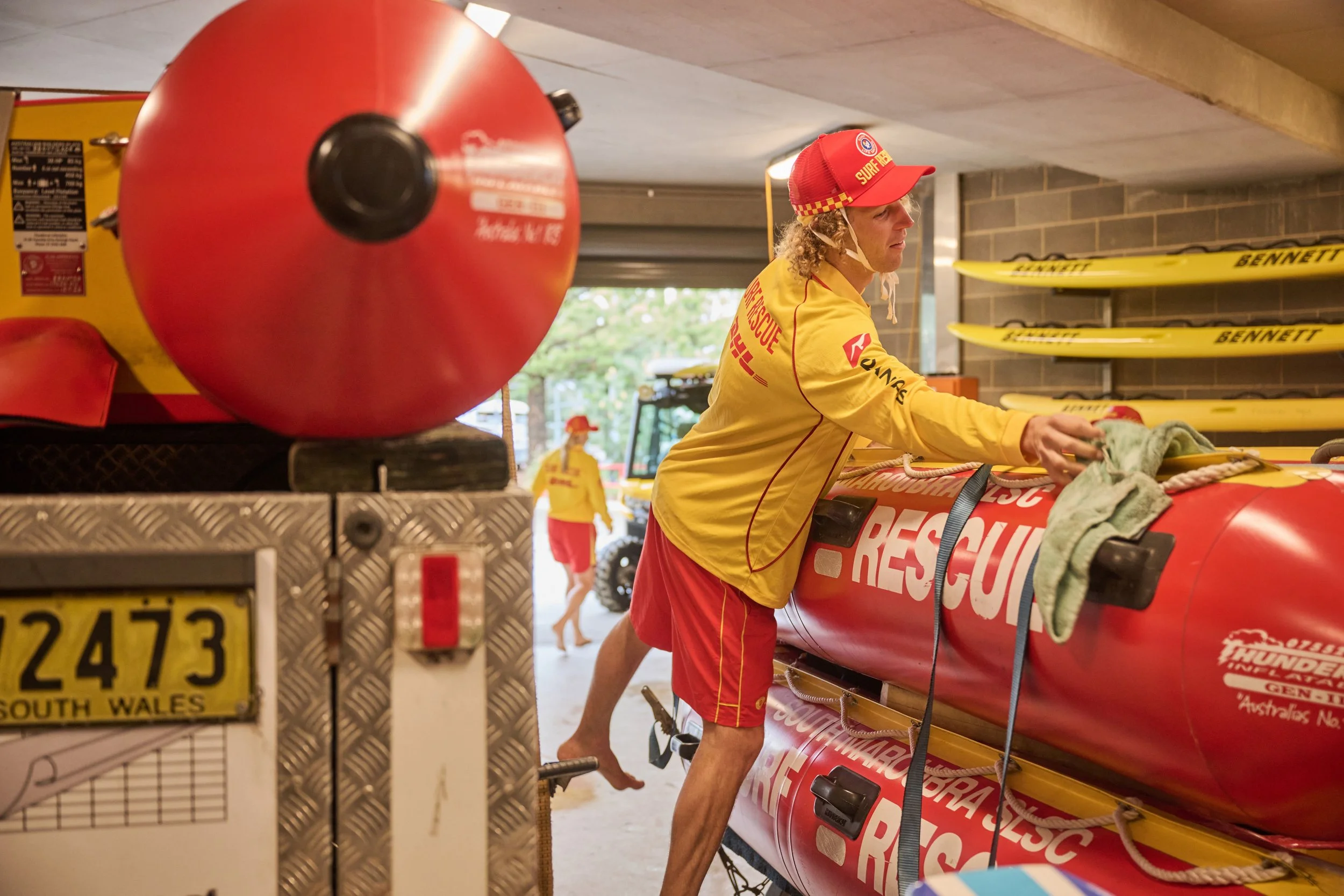 A member of a rescue team wearing a yellow shirt and red cap is working with equipment in a storage area, with other rescue personnel in the background.