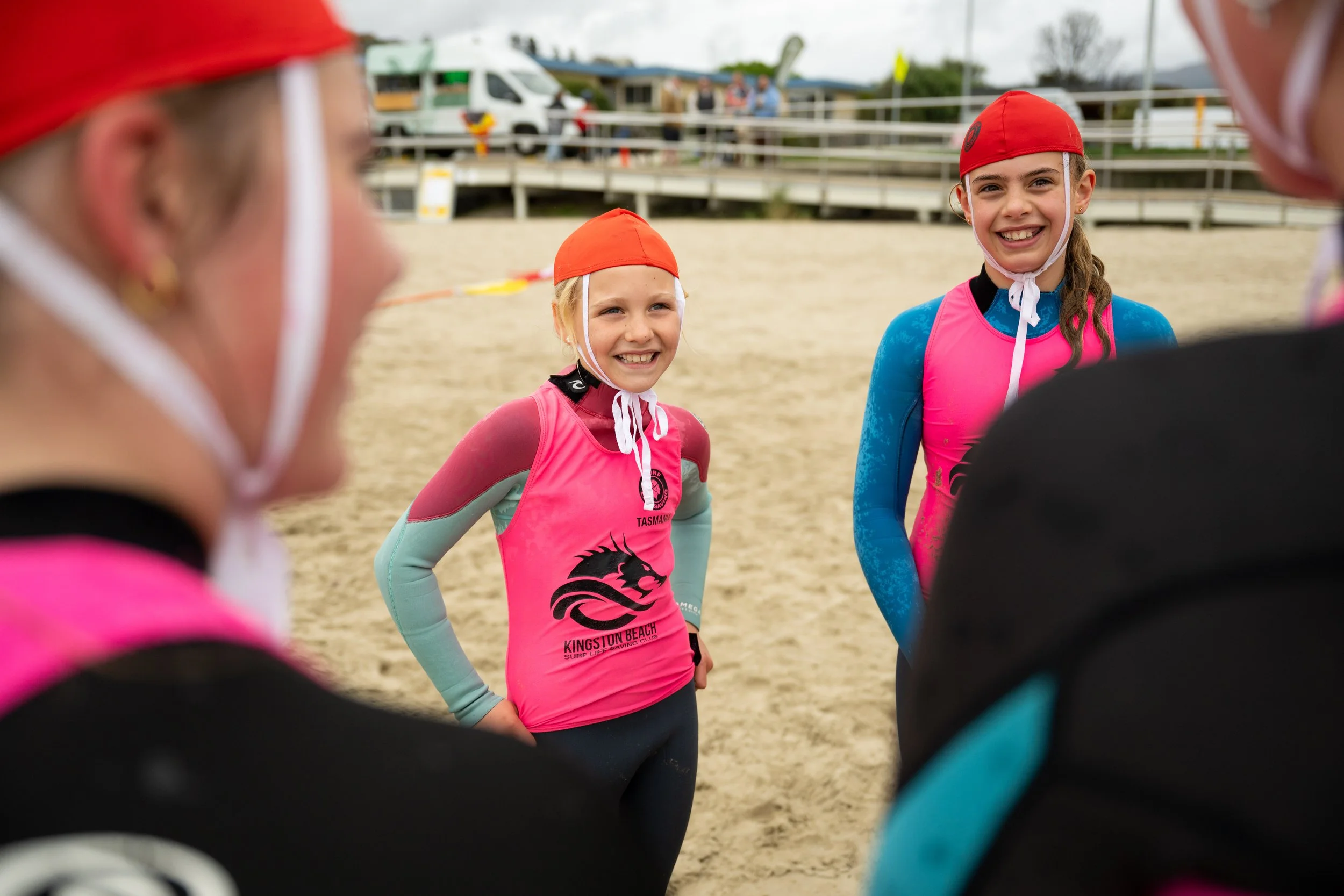 Young girls wearing wetsuits and red swim caps standing on a beach, having a conversation with two adults.