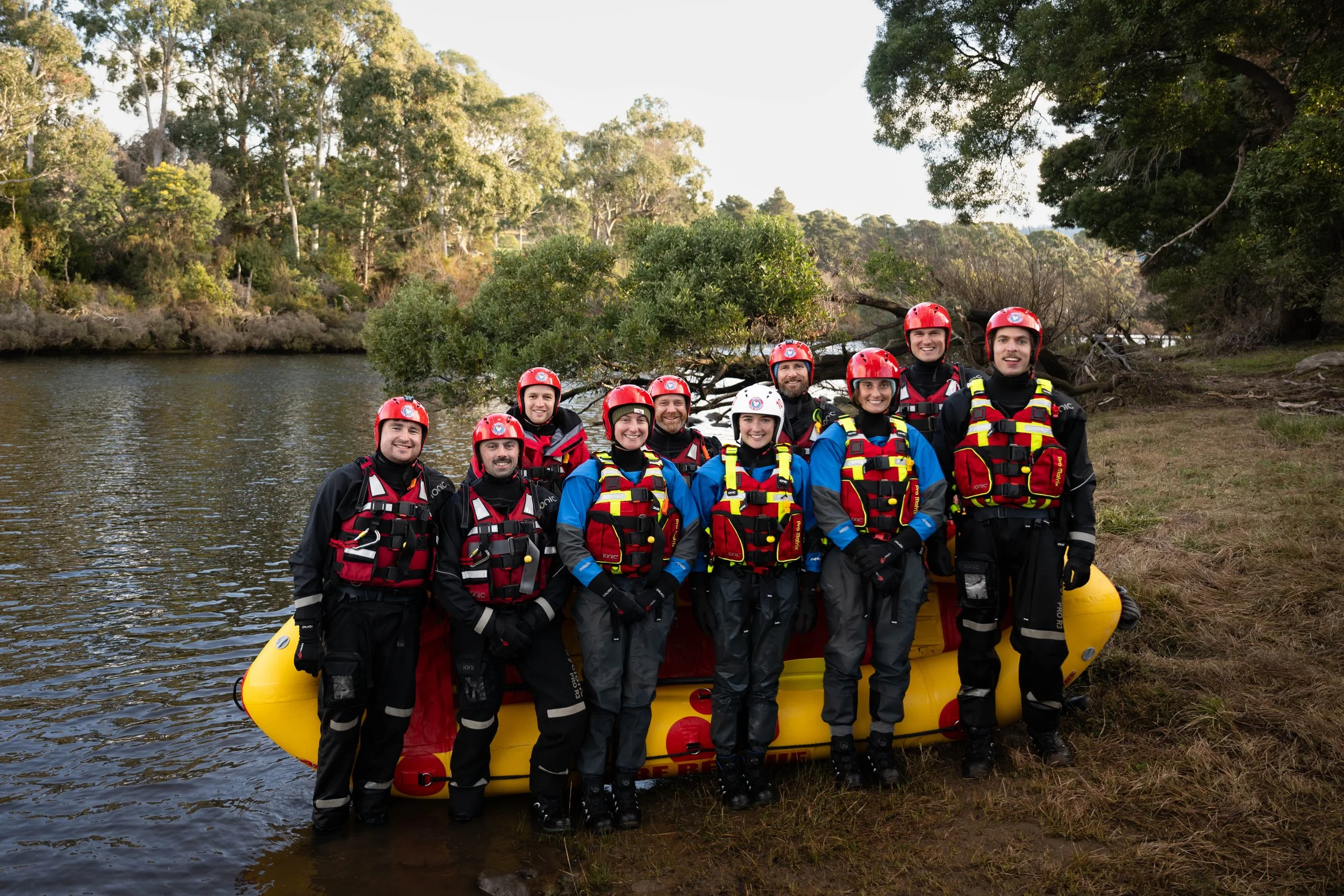 Group of nine people in swift water rescue gear standing in front of a yellow rescue boat by a river, surrounded by trees.
