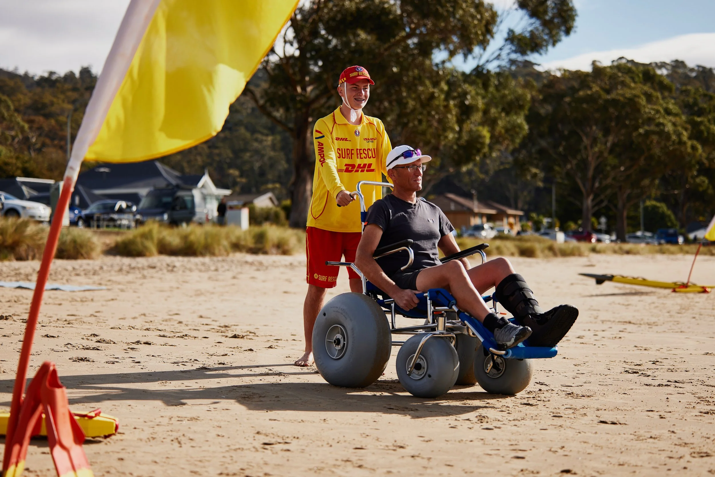 A person in a wheelchair on a beach with a surf rescue worker standing behind them. The rescue worker is smiling and dressed in a yellow surf rescue uniform. There is a large yellow flag in the foreground and trees and parked cars in the background.