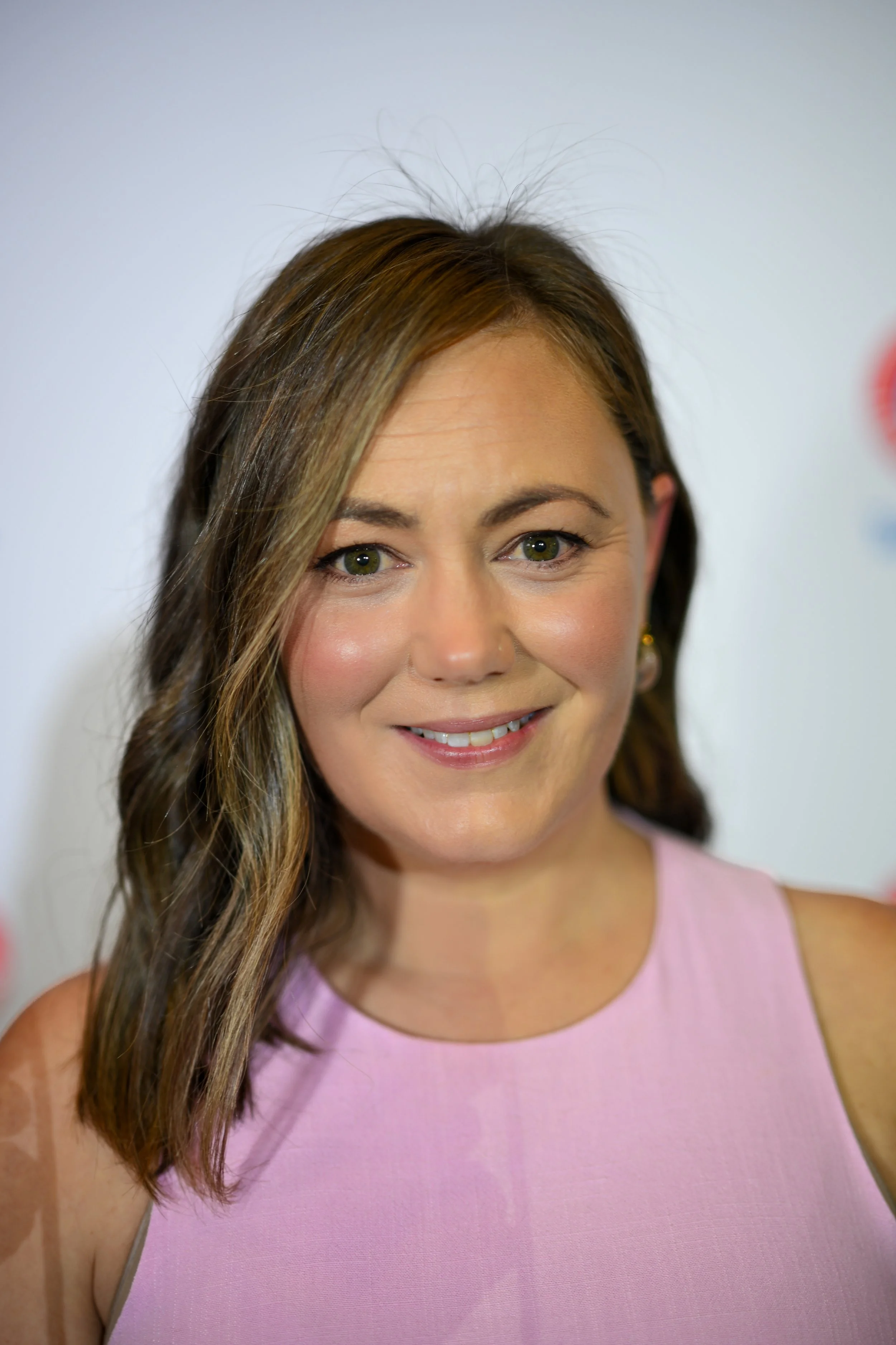 Close-up of a woman smiling, wearing a pink sleeveless top, with brown hair and green eyes, against a plain white background.