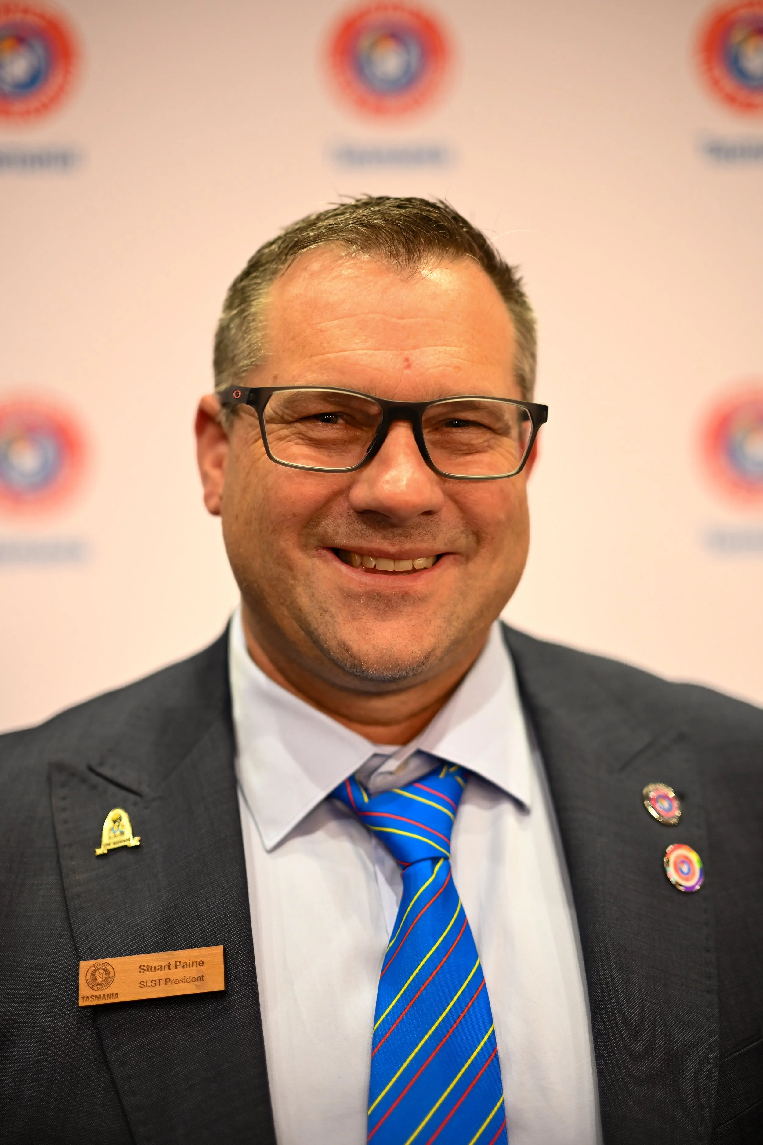 A smiling man wearing glasses, a dark suit, a white shirt, and a blue striped tie. He has short hair and is wearing lapel pins and badges, including a name tag that reads 'Stuart Paine, SLST President, Tasmania.' The background features a white backdrop with logo patterns.