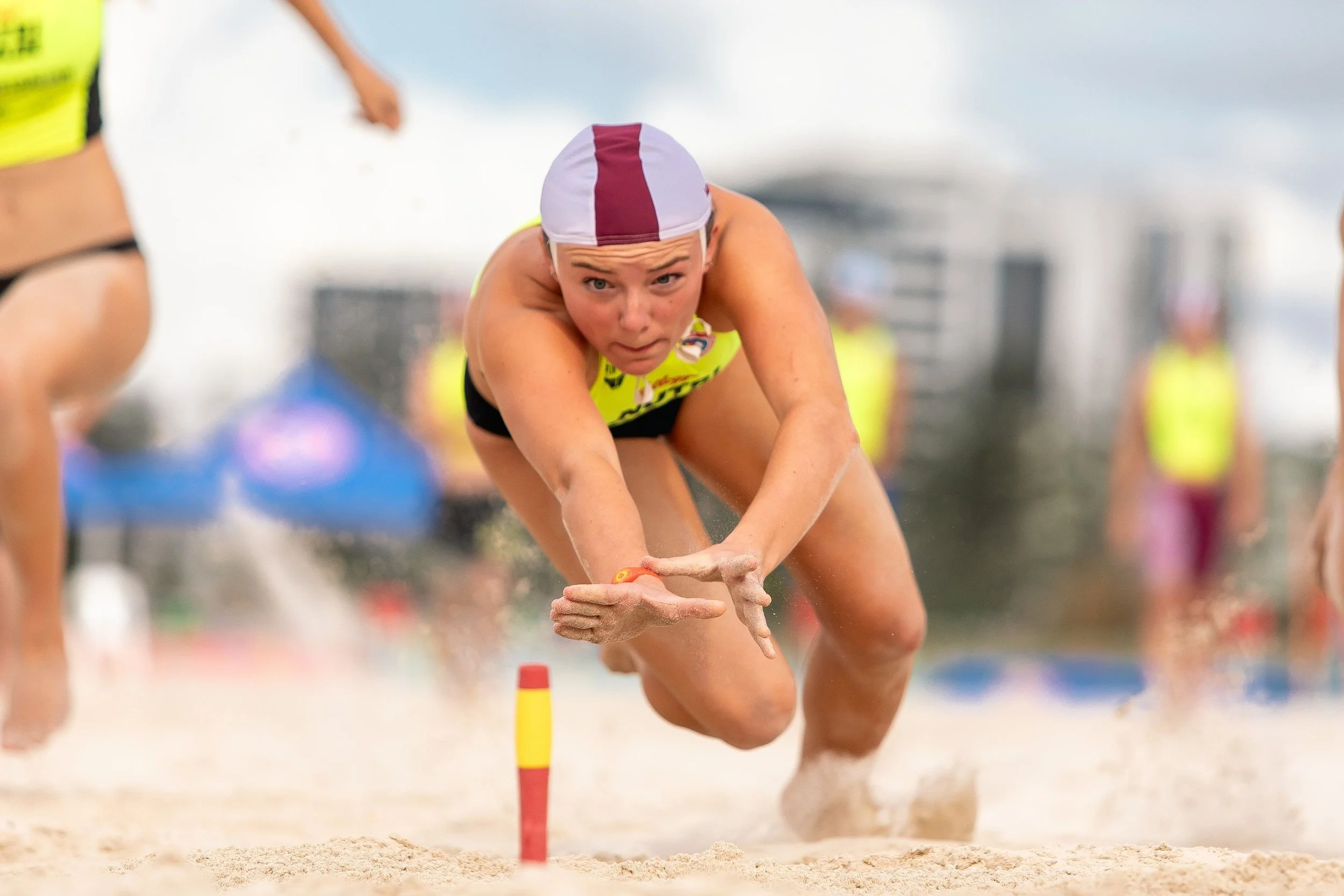 Female beach volleyball player diving in sand, wearing a white and maroon cap and neon yellow sportswear, during a match with other players in the background.