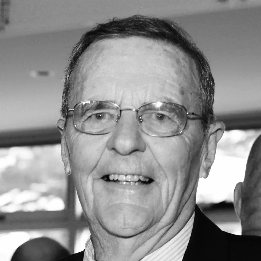 Black and white photo of an elderly man with glasses, smiling, wearing a suit, and with windows in the background.