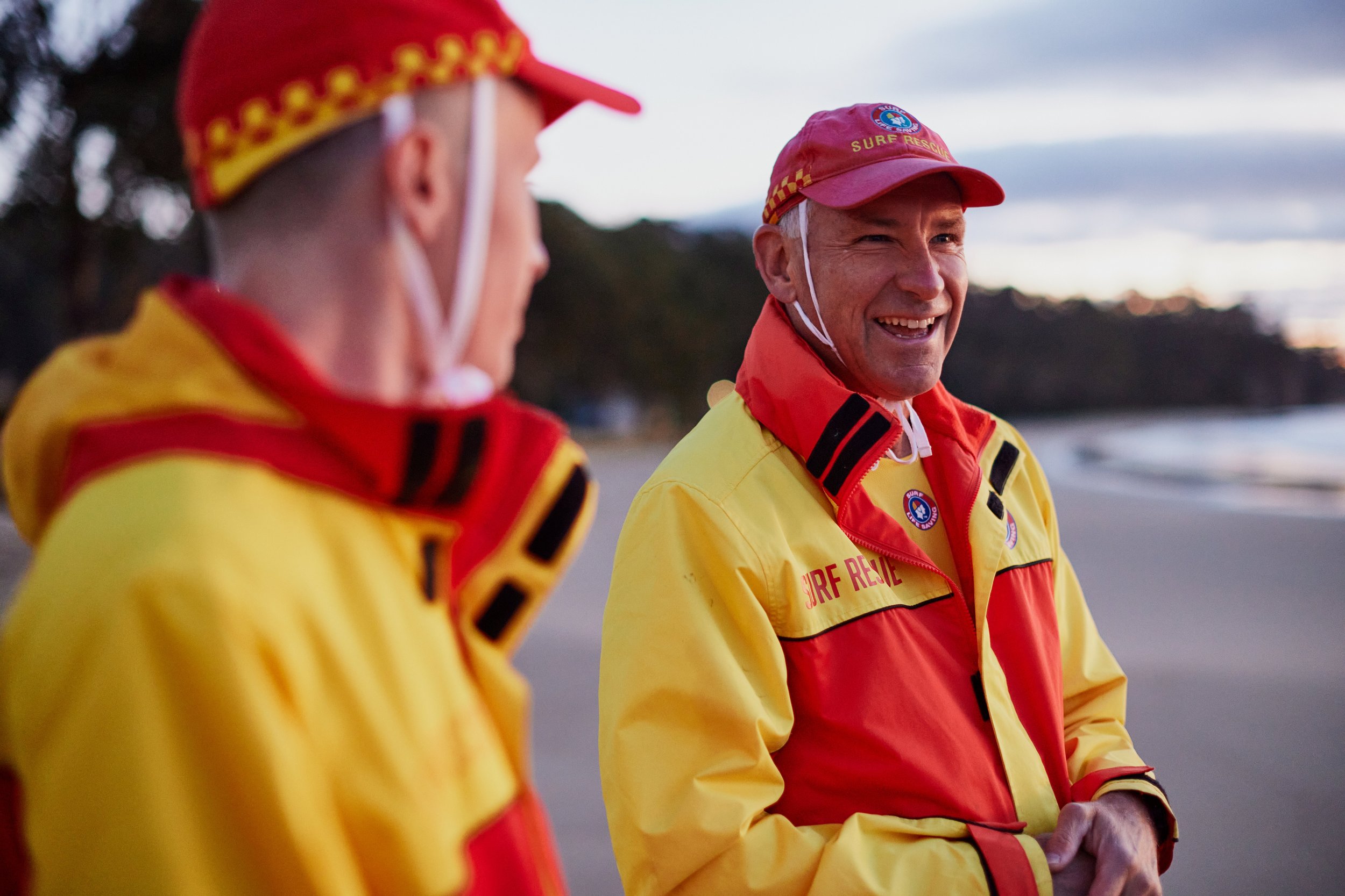 Two people wearing yellow and red surf rescue uniforms and red caps having a conversation outdoors at sunset.