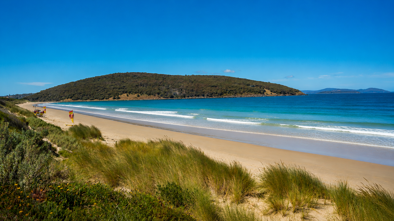 A sandy beach with grassy dunes in the foreground, colorful umbrellas and lounge chairs along the shoreline, calm blue ocean waves, and a green hilly island in the distance under a clear blue sky.