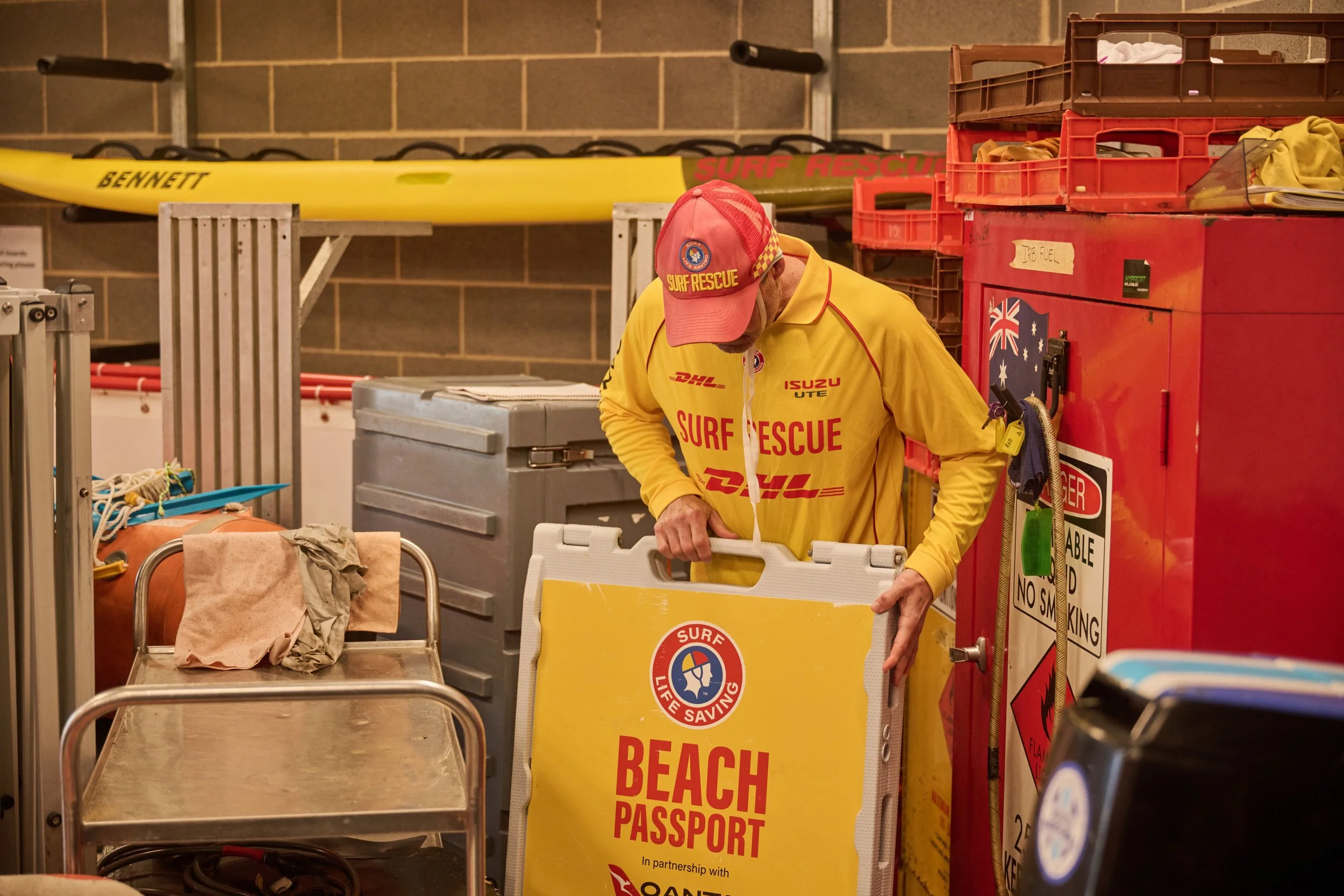 A person dressed in yellow surf rescue attire is working with a yellow emergency rescue box in an indoor storage area. There are surf rescue boards, crates, and various emergency supplies around.