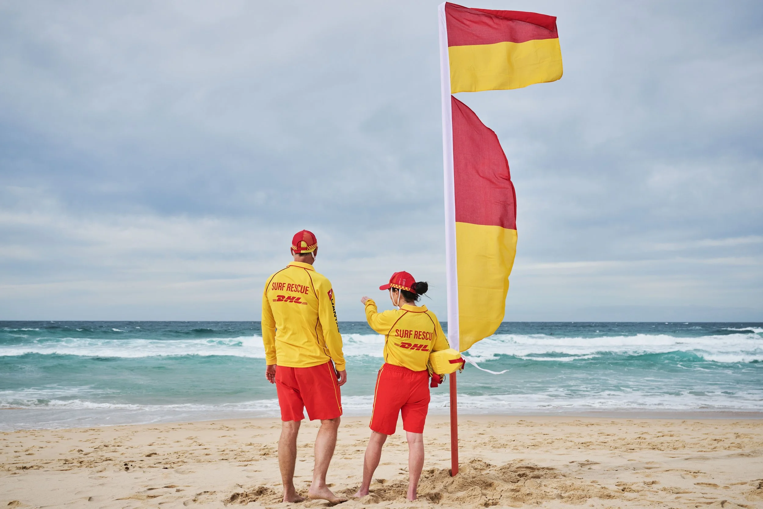 Two surf rescue team members in yellow and red uniforms standing on a beach, facing the ocean, with large red and yellow rescue flag nearby, under cloudy sky.