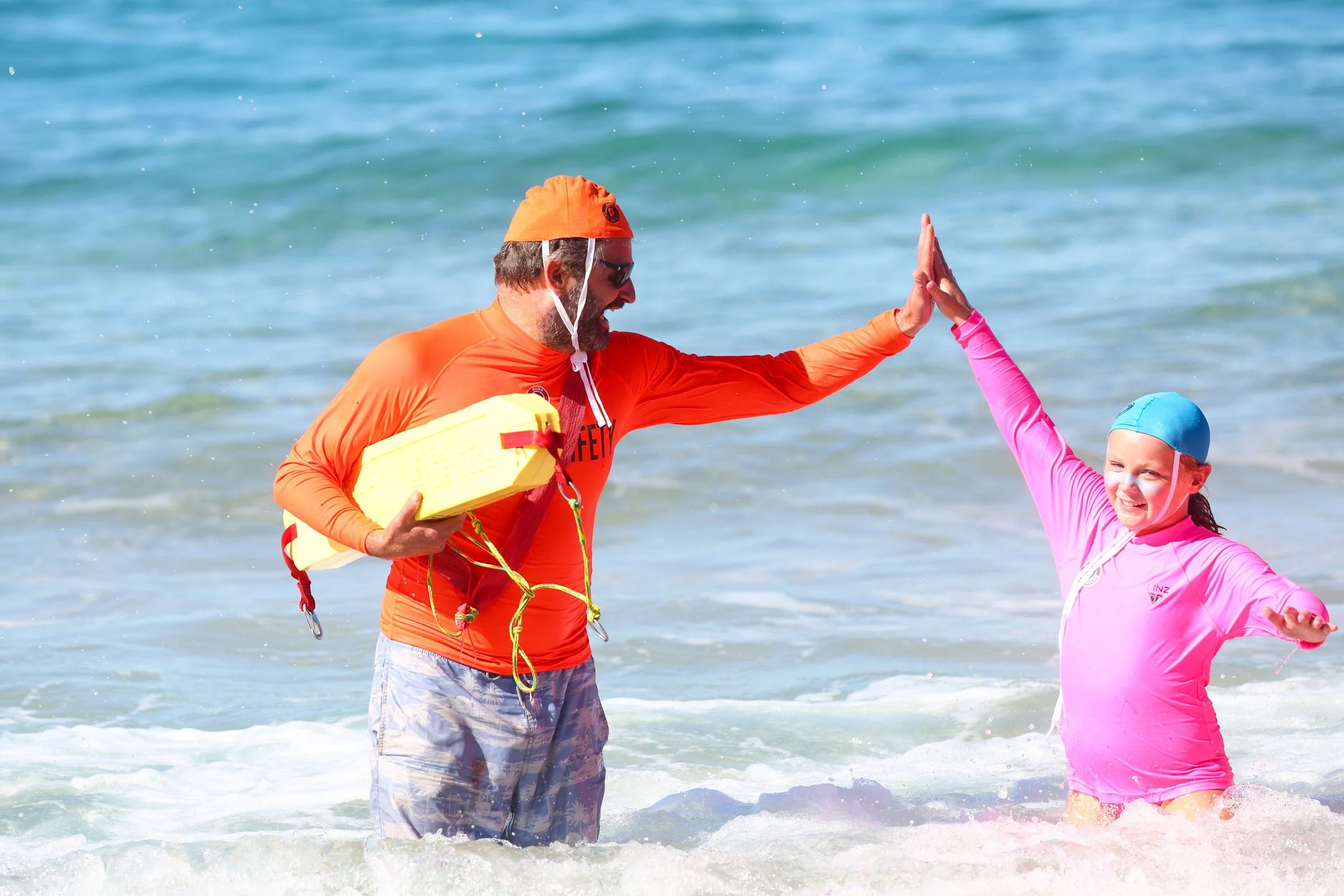 A man and a girl wearing life jackets and swim caps enjoying a high five in the ocean water.