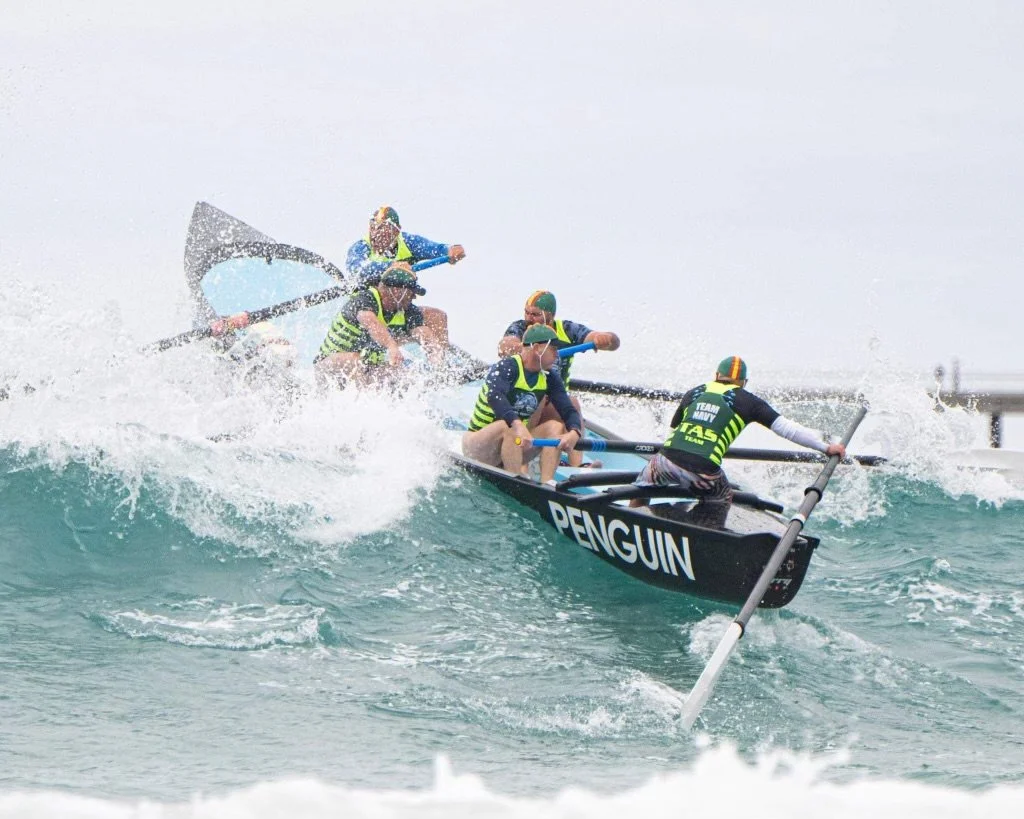 A team of six people in a boat labeled 'PENGUIN' racing on a large ocean wave, using paddles to control the boat.