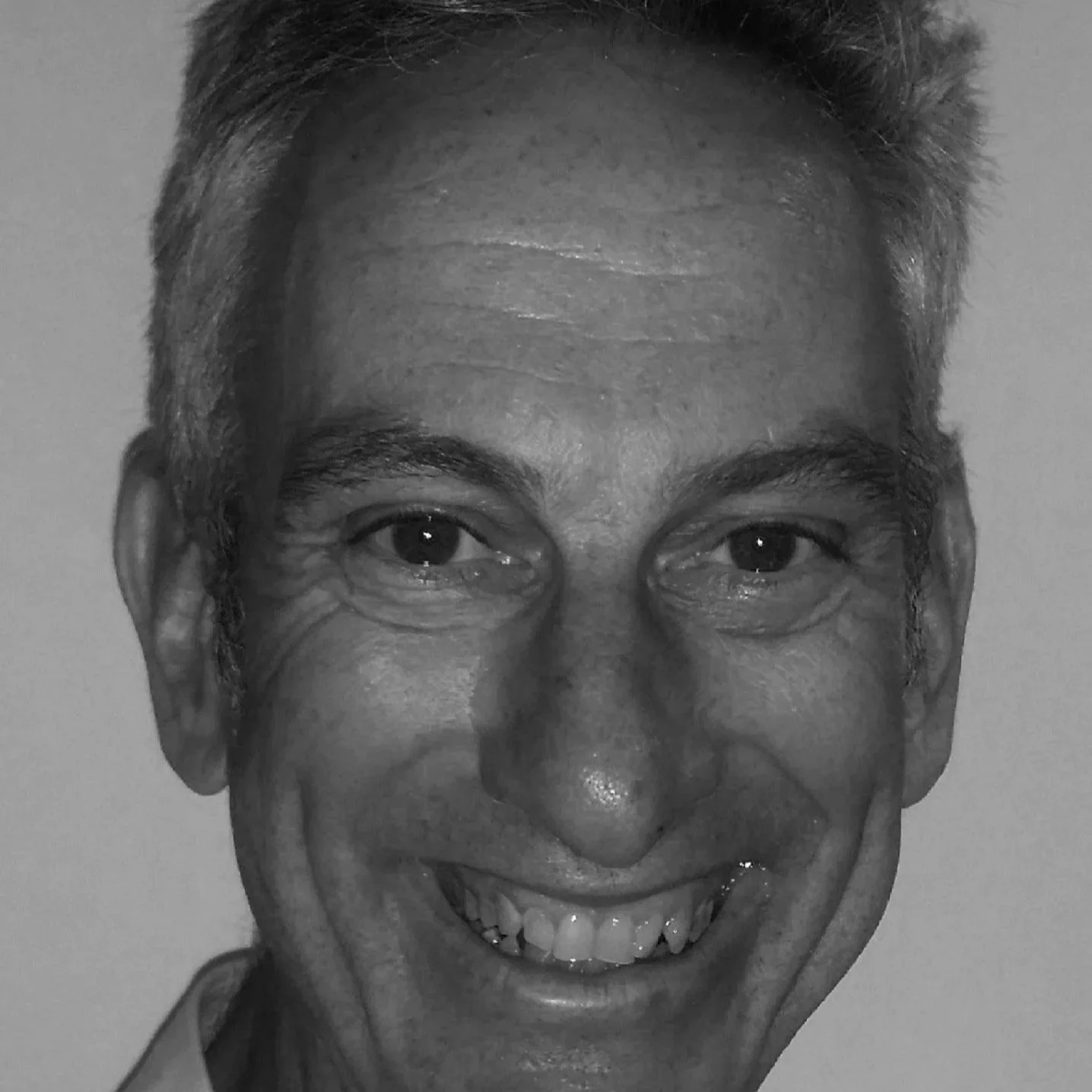 A black-and-white close-up of a smiling elderly man with short hair, smiling at the camera.