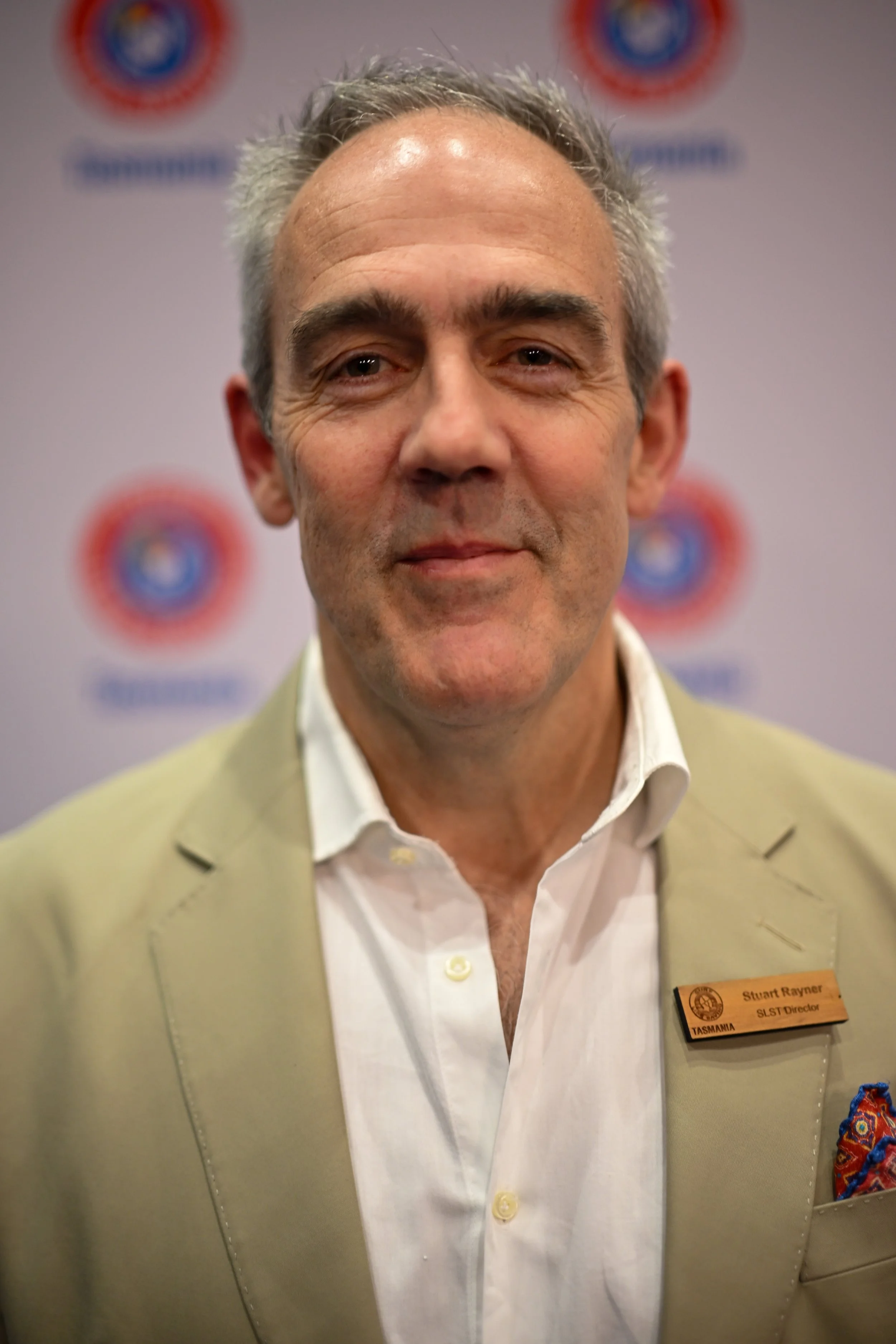 Close-up photo of a smiling middle-aged man with gray hair, wearing a cream-colored suit jacket and white dress shirt. He has a name tag that reads "Stuart Rayner" and "Tasmania SLT Director." The background shows a blurred logo or emblem.