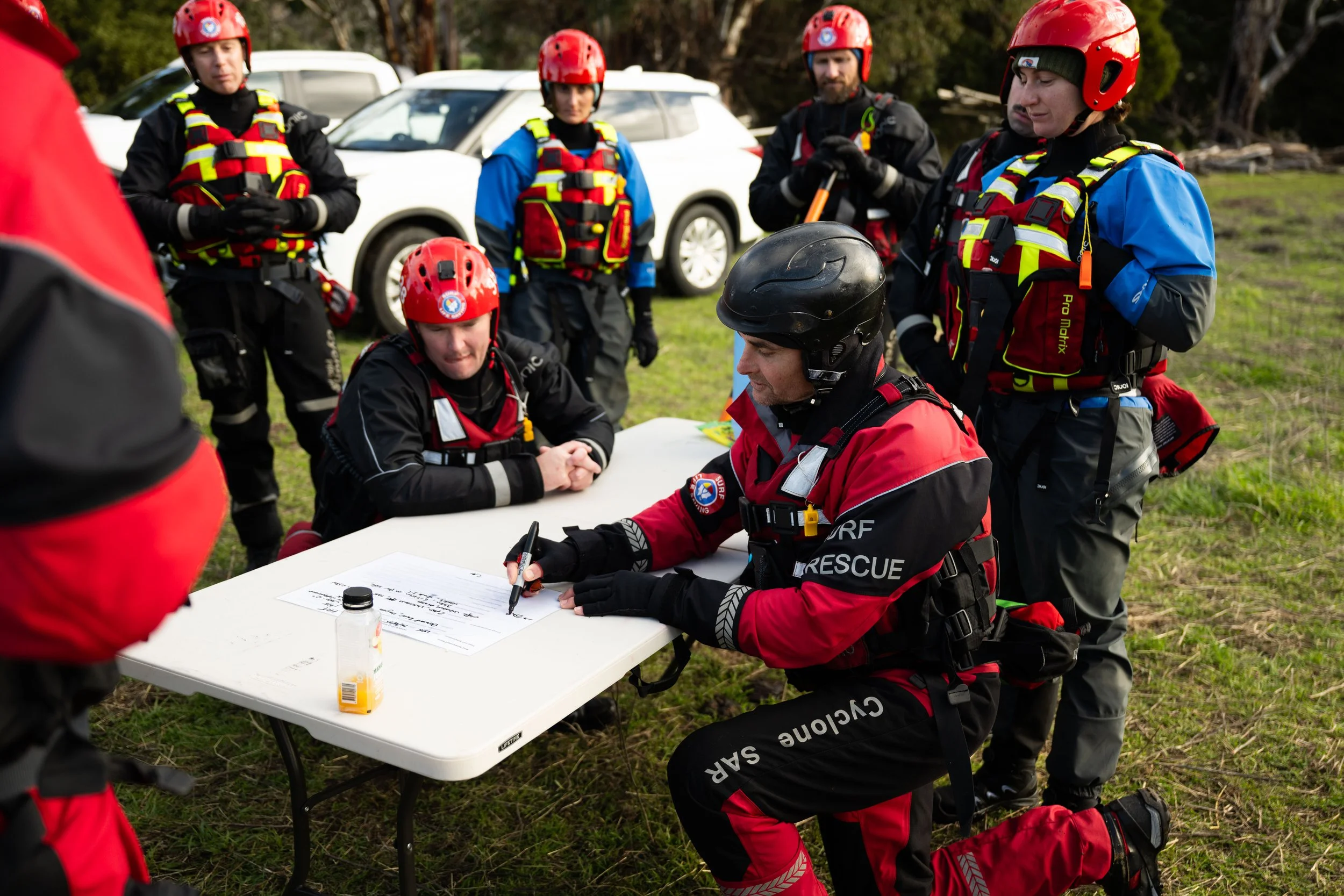 Rescue team members wearing helmets and safety gear gathered around a table outdoors, reviewing documents as part of a rescue operation.