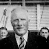 Black and white photo of a man with white hair, wearing a suit and tie, standing outdoors with several people in the background.