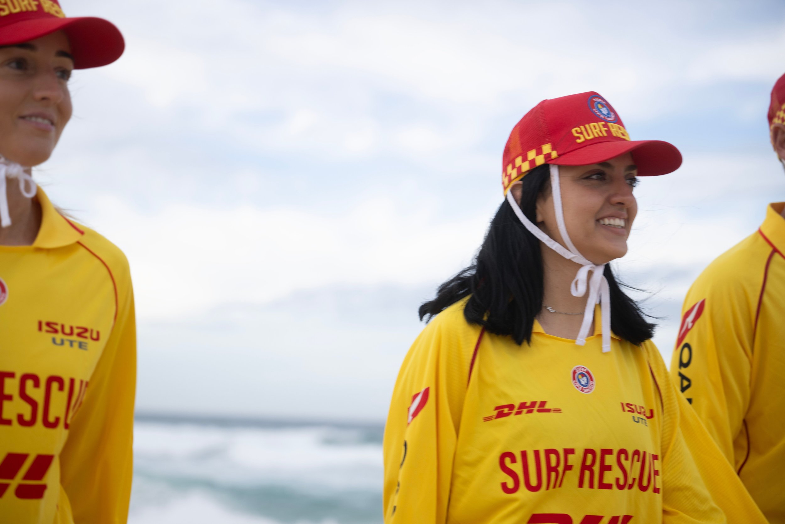 Three people wearing yellow surf rescue jackets and red caps standing outdoors against a cloudy sky.