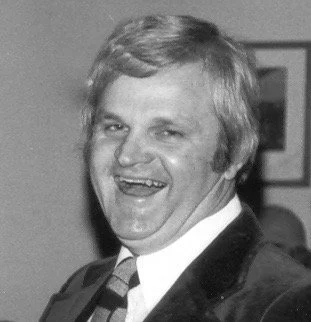 A black-and-white photo of a smiling man with light-colored hair, wearing a suit and tie, in an indoor setting.