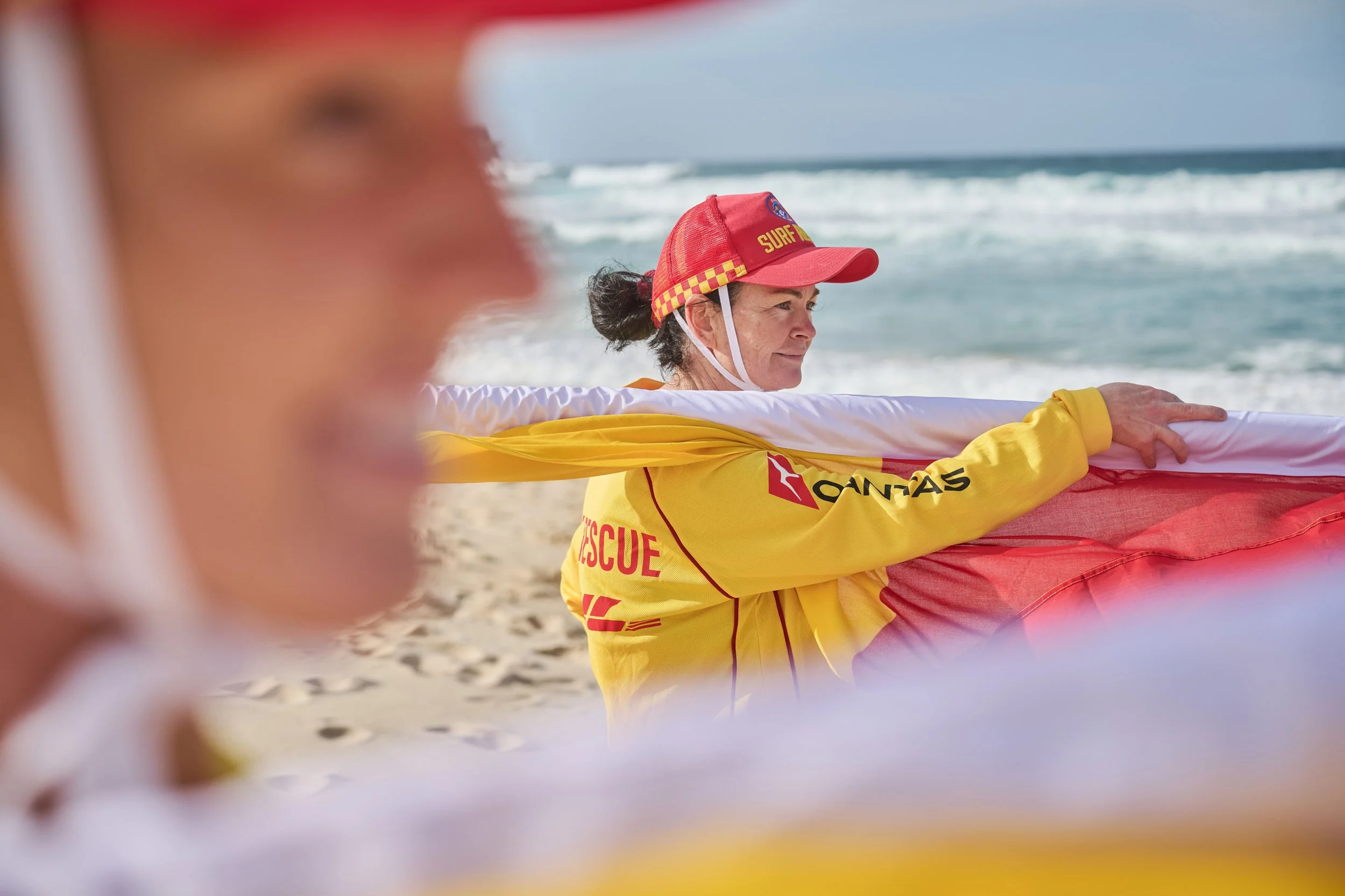A female surf rescue worker on the beach holding a red rescue tube, wearing a yellow rescue uniform and a red cap with the word 'SURF' on it, with ocean waves in the background.