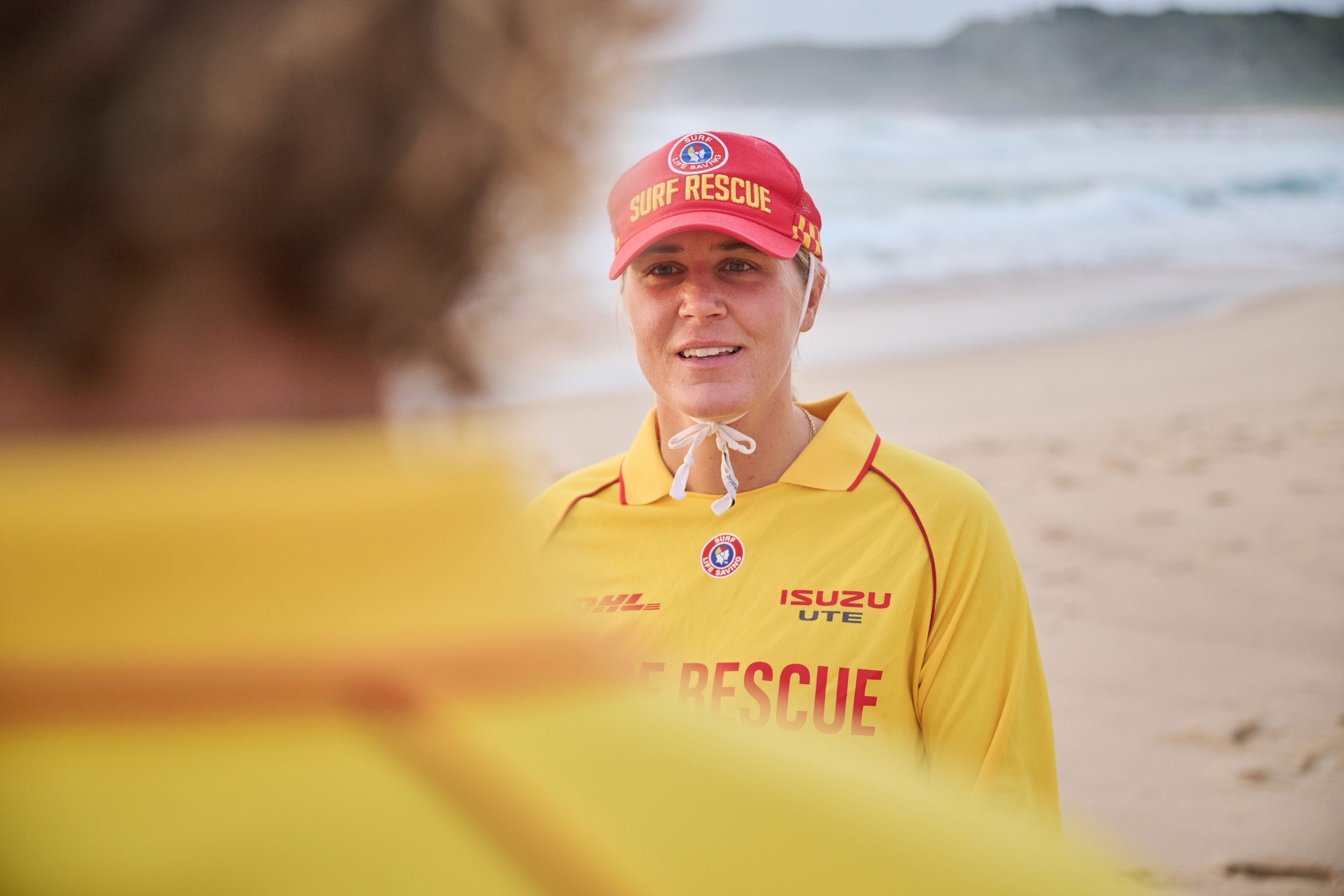 A female surf rescue worker with fair skin and blonde hair, wearing a red cap and yellow rescue uniform, standing on a beach near the ocean, engaged in a conversation.
