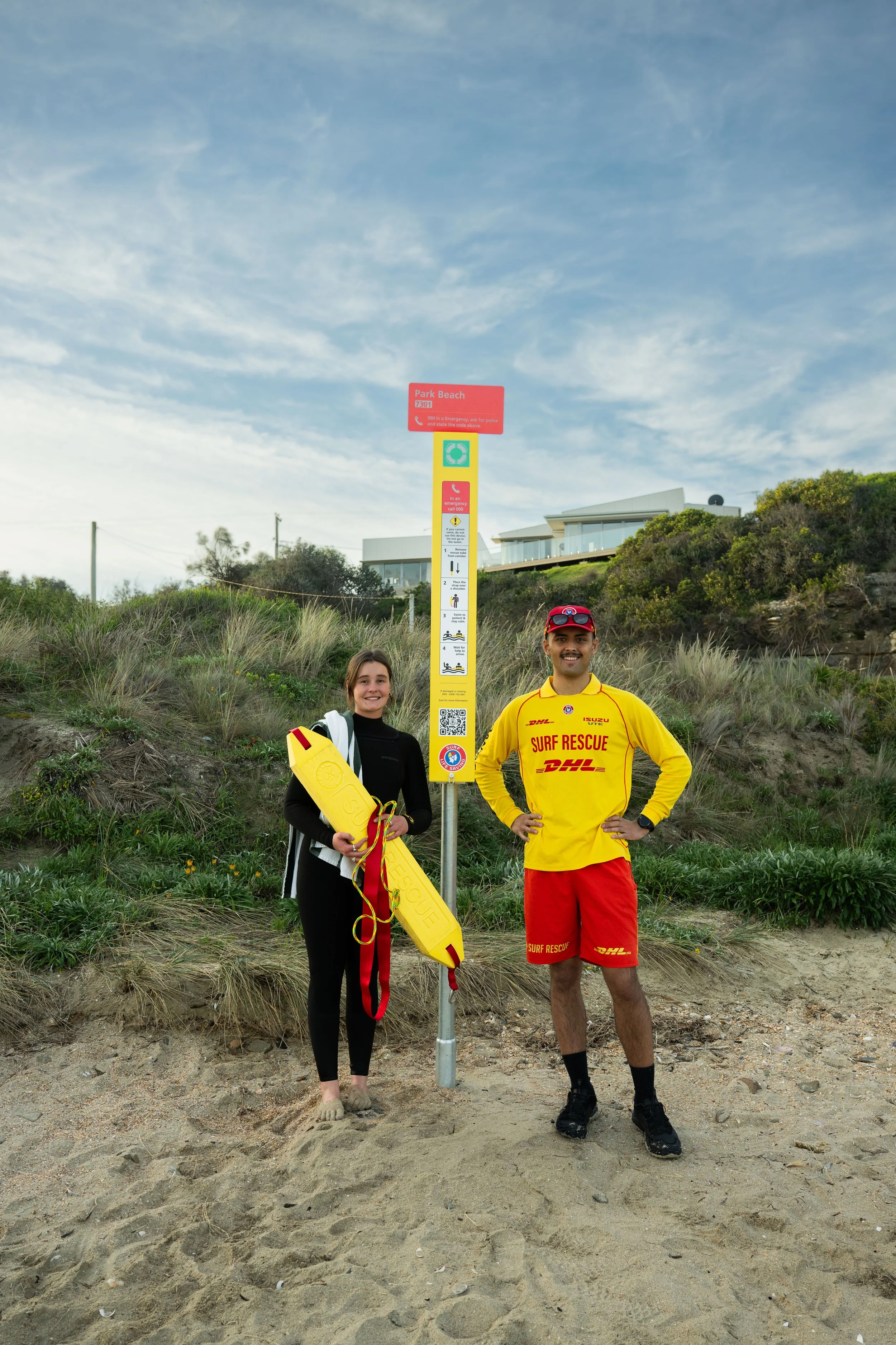 A female and male surf rescue team members standing on the beach next to a yellow rescue pole and emergency signs, smiling at the camera with buildings and greenery in the background.